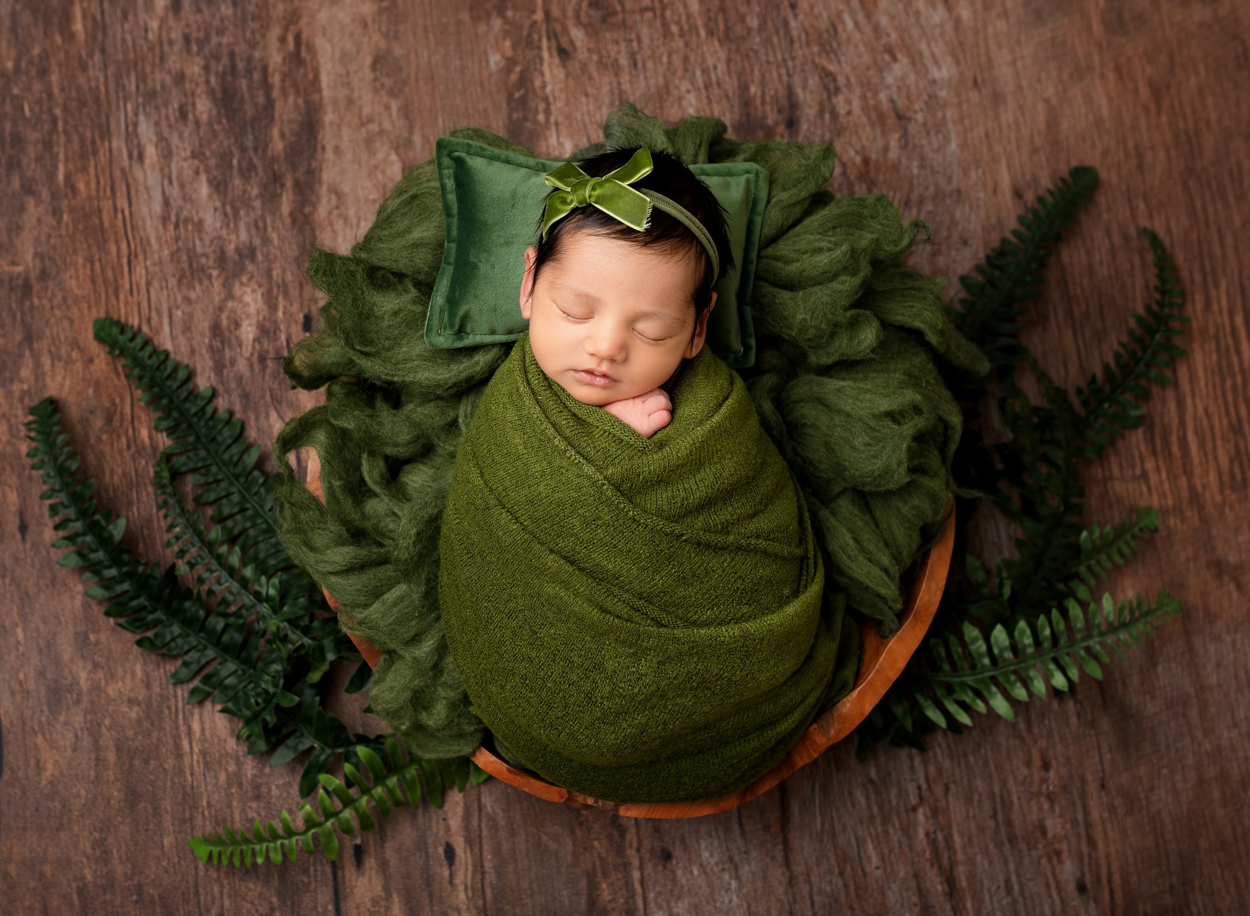 Sleeping newborn baby wrapped in green swaddle, lying on green and brown pillows surrounded by ferns on a wooden floor.