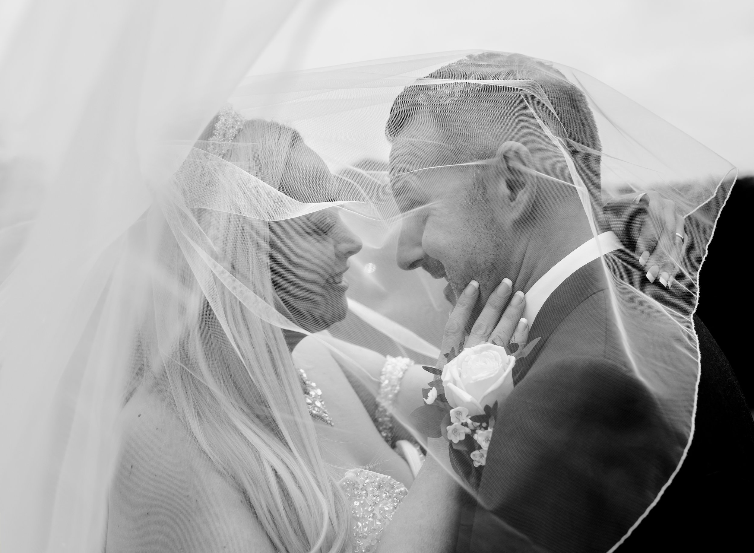 Black and white photo of a bride and groom under a veil, smiling and touching faces during their wedding.