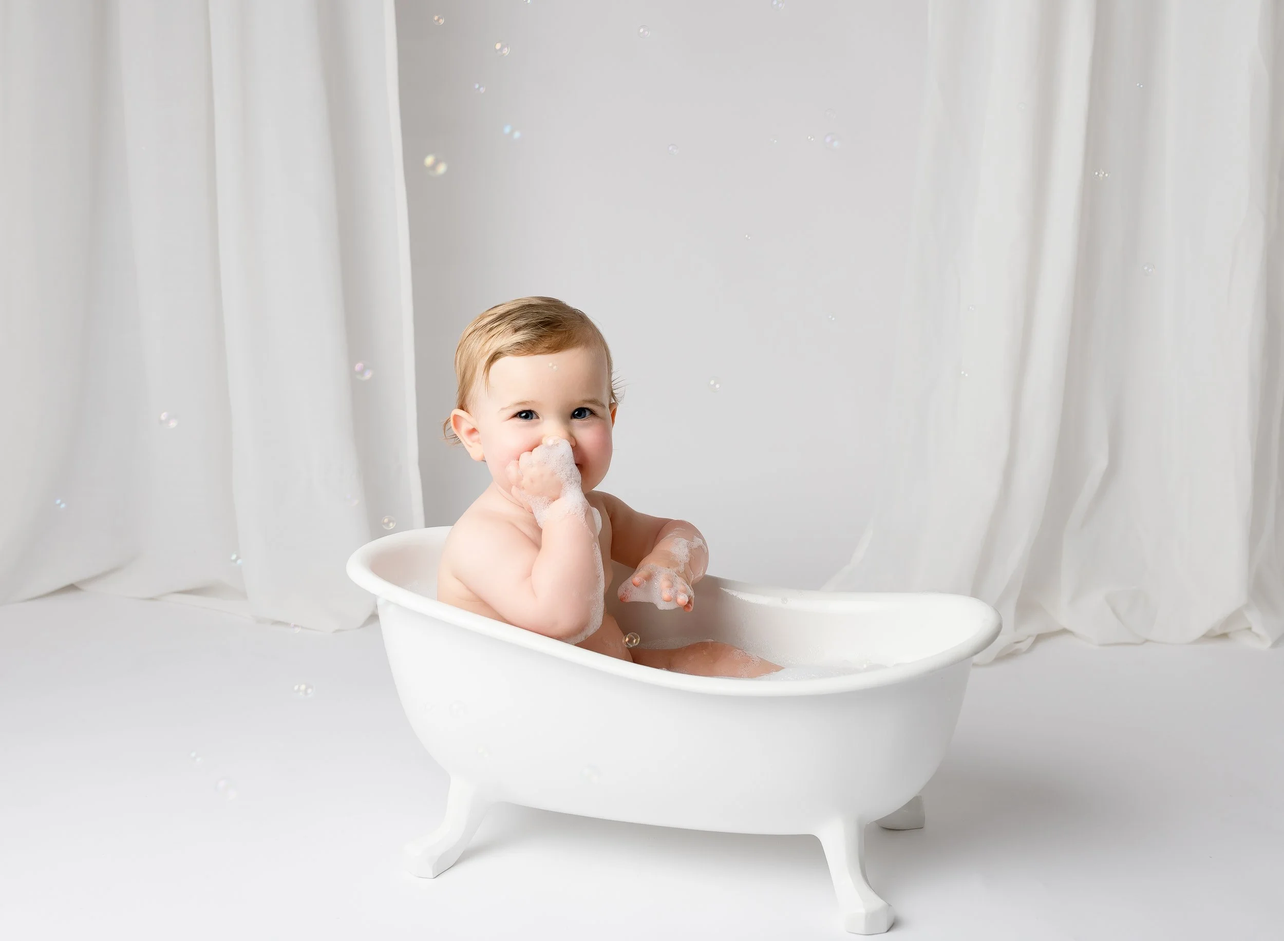 Baby taking a bath in a small white bathtub with soap bubbles, smiling and looking at the camera.