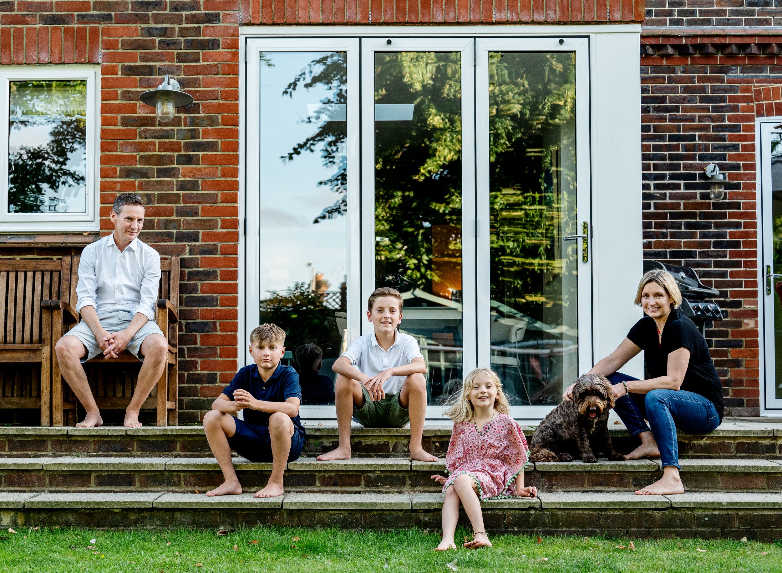 Family sitting on outdoor steps in front of a brick house, including two adults, three children, and a dog.