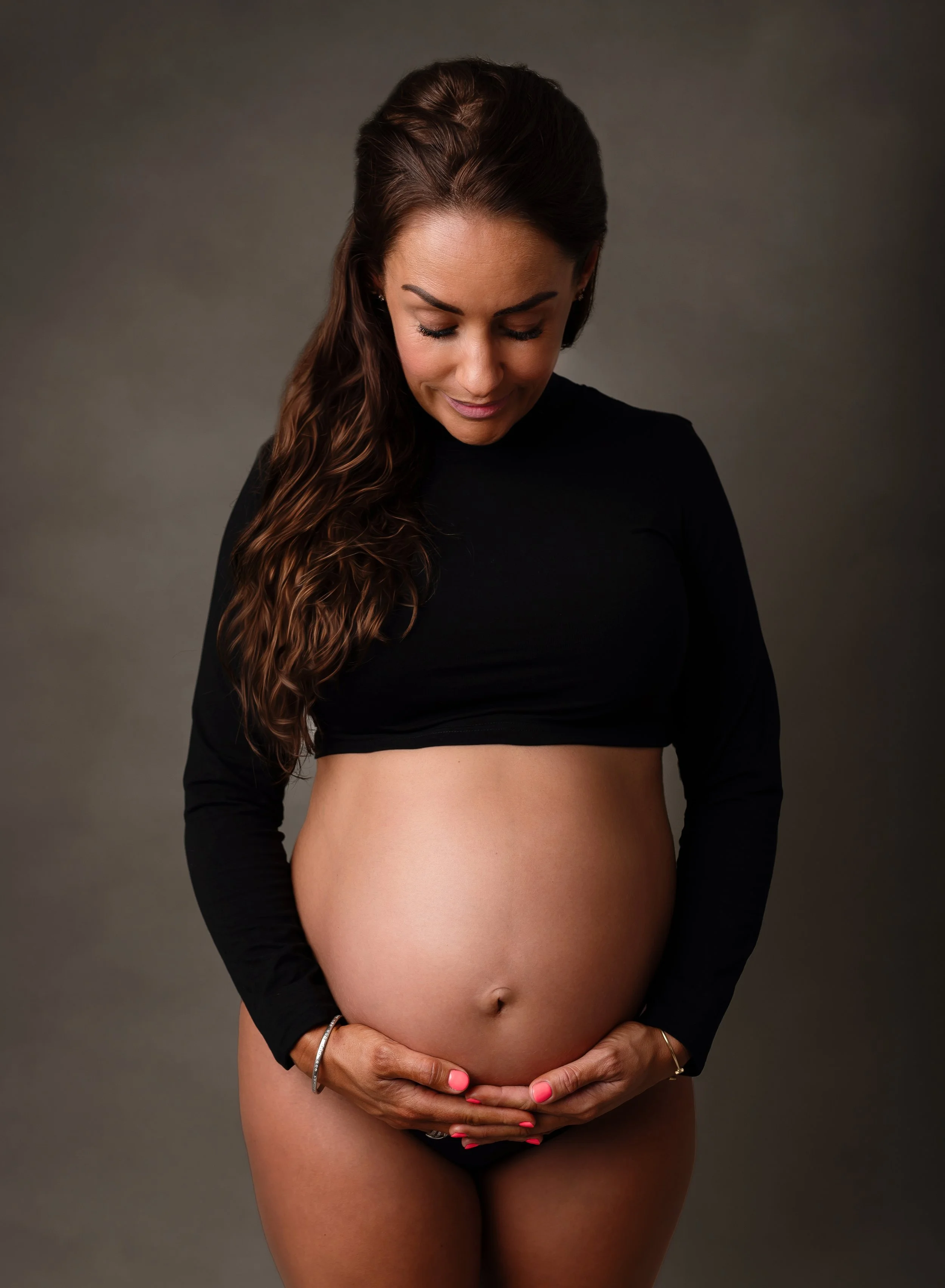 A pregnant woman with long brown hair, wearing a black long-sleeve top, is smiling gently and holding her pregnant belly with both hands, looking down at it against a plain gray background.