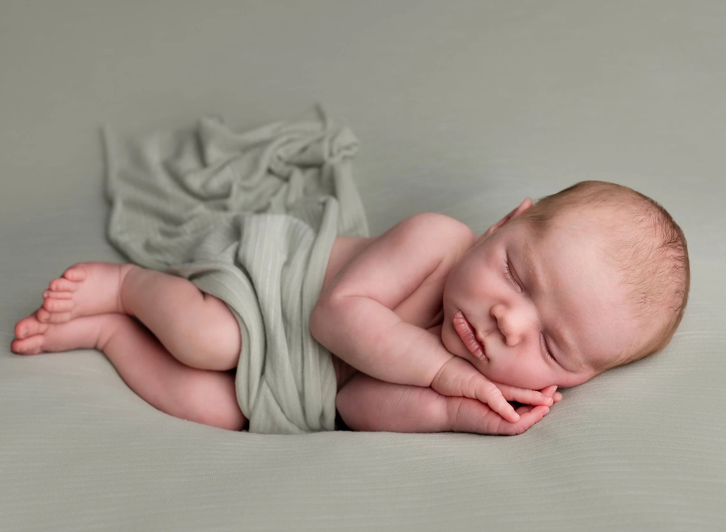 A sleeping baby lying on a soft surface, curled up with hands near face, wrapped in a light-colored cloth.