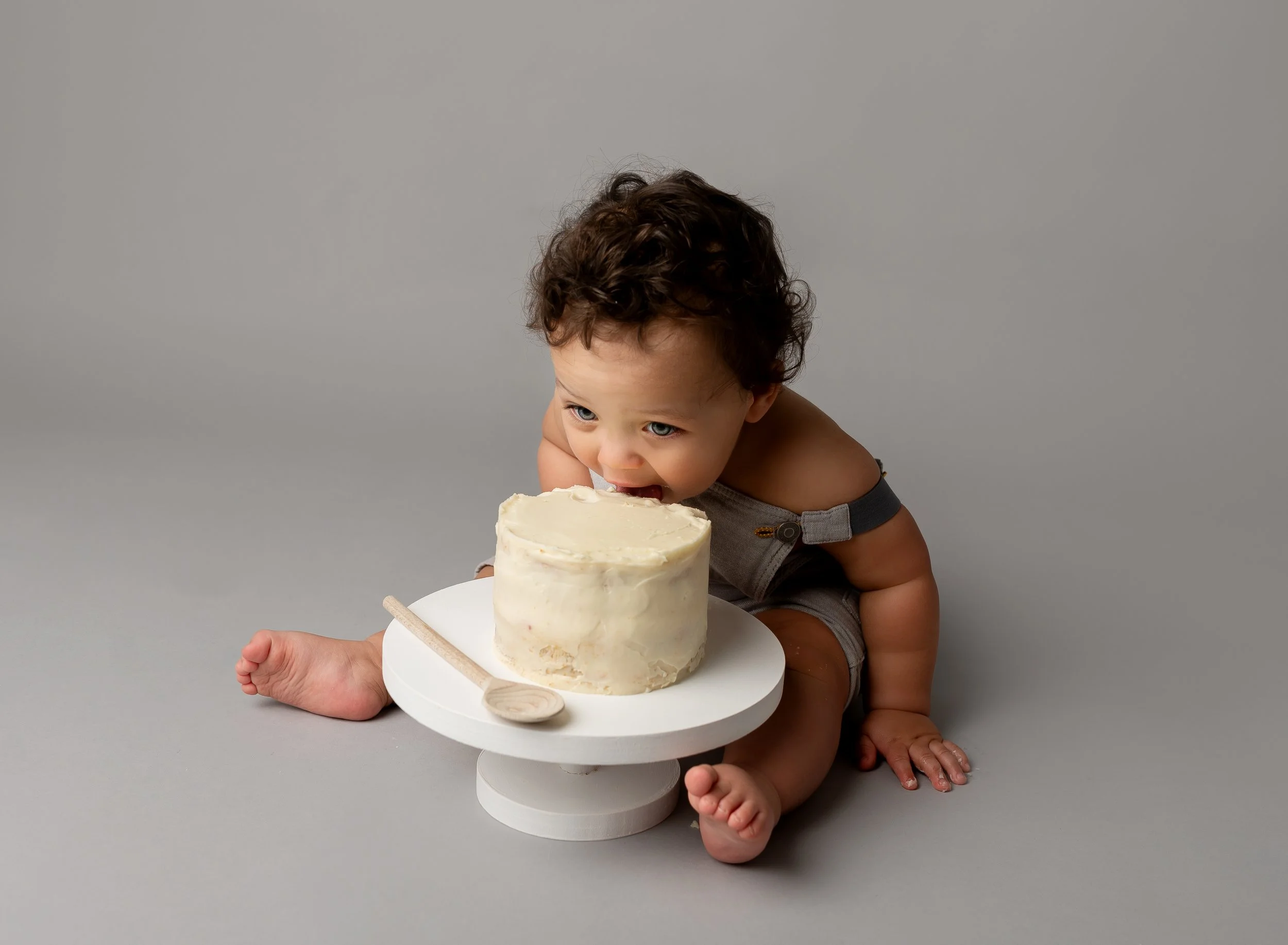 A young child with curly hair, sitting on a gray floor, is licking a white frosted cake with white frosting and a wooden spoon placed on a white cake stand.