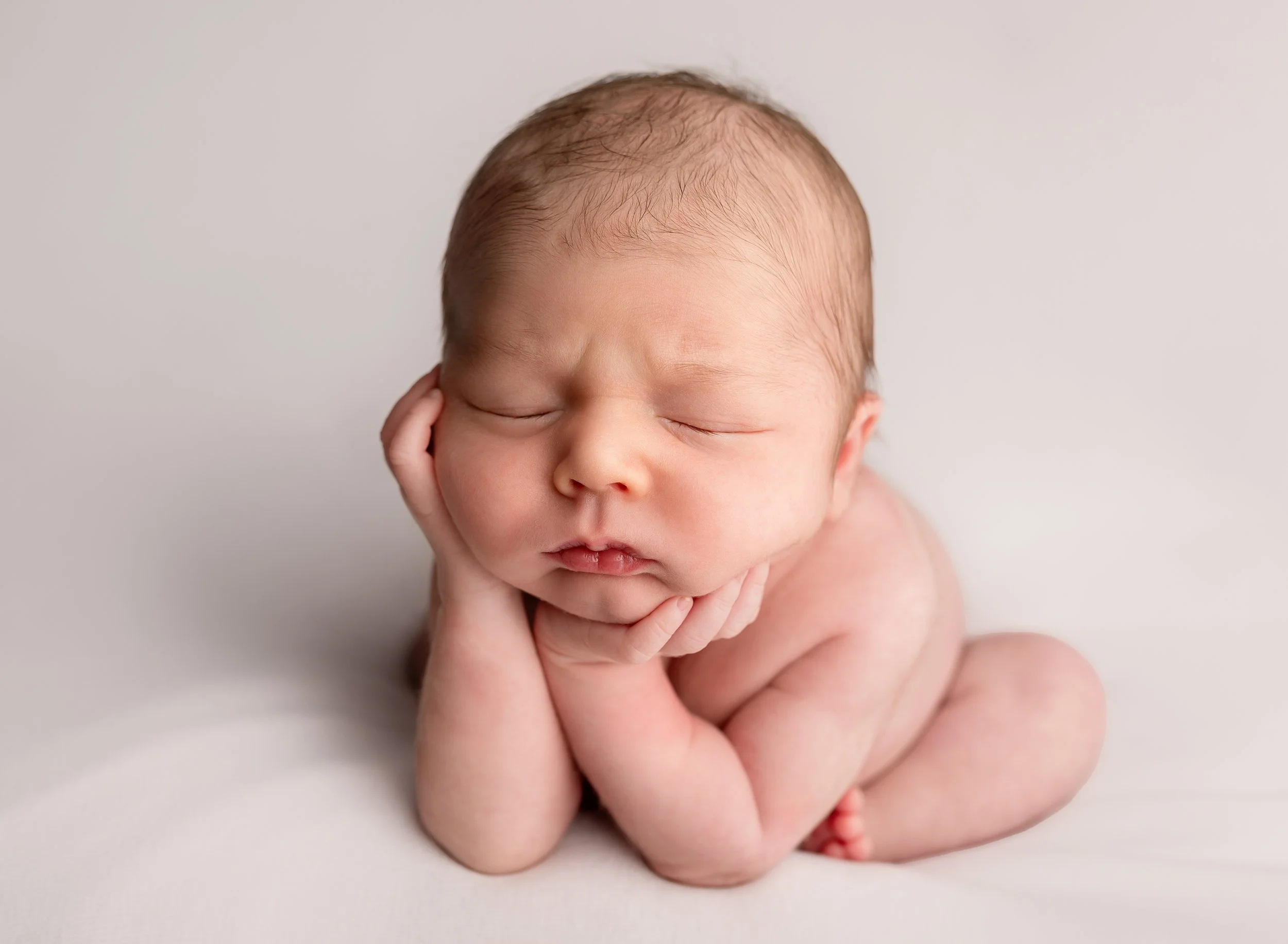 Close-up of a sleeping newborn baby with hands resting on cheeks, on a plain white background.