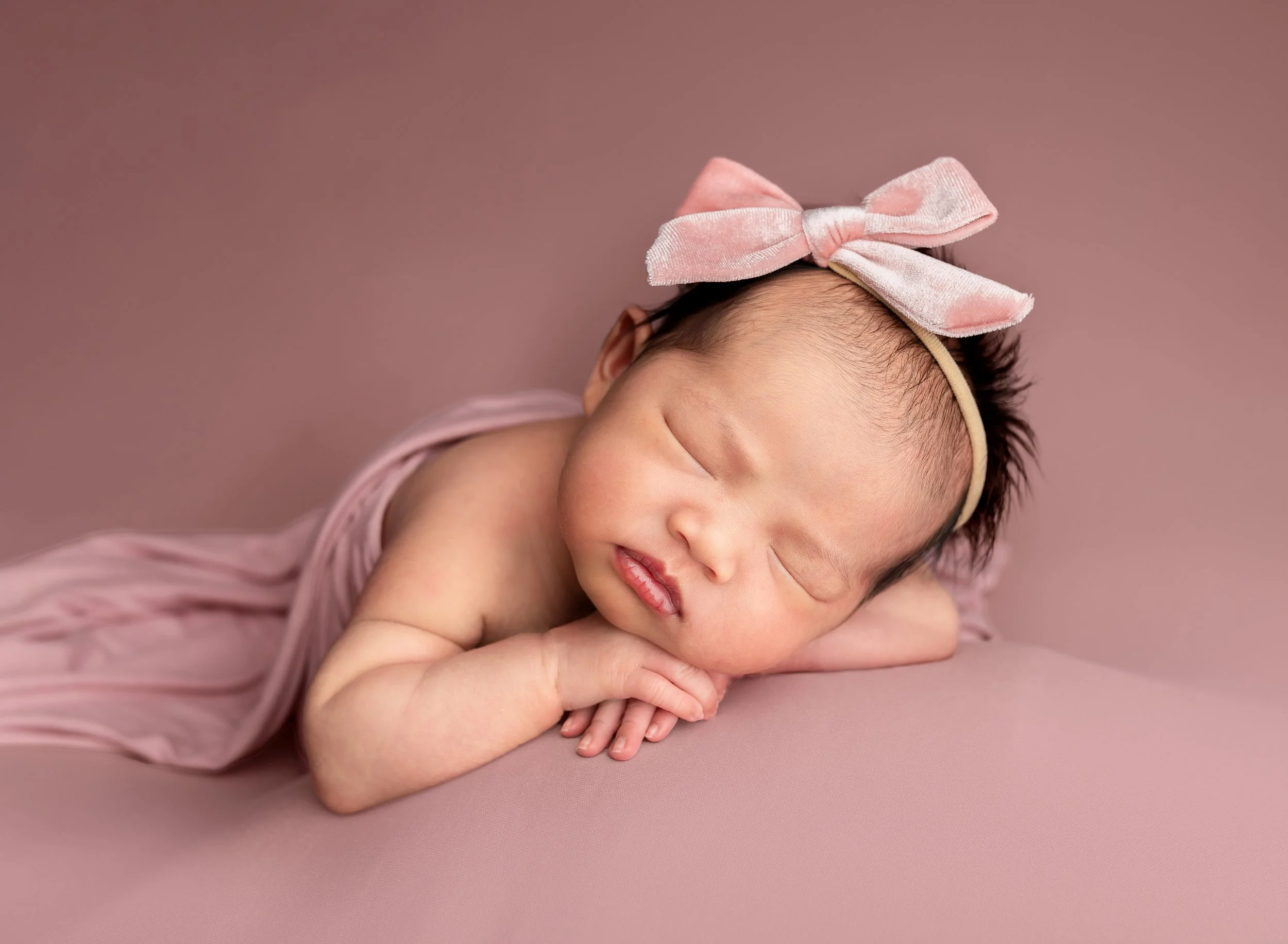 A sleeping baby girl with a pink bow headband, lying on her side on a pink surface, dressed in a light pink outfit.