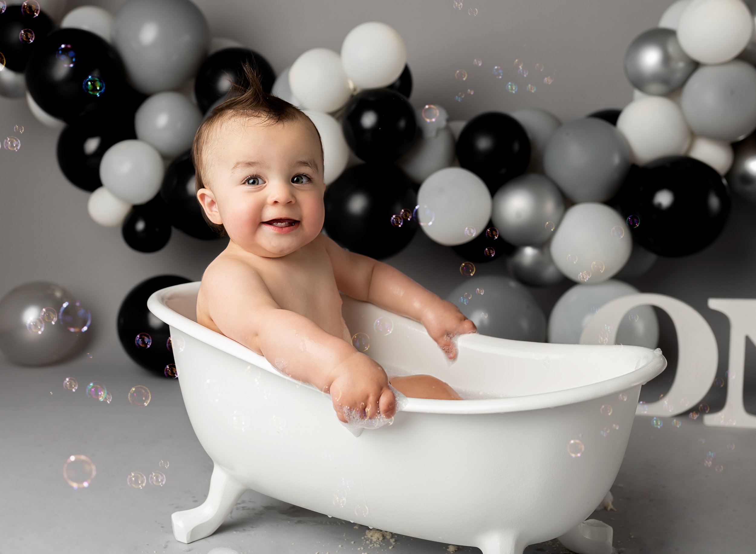 A smiling baby sitting in a small white bathtub with black, white, gray, and silver balloons in the background, surrounded by soap bubbles.