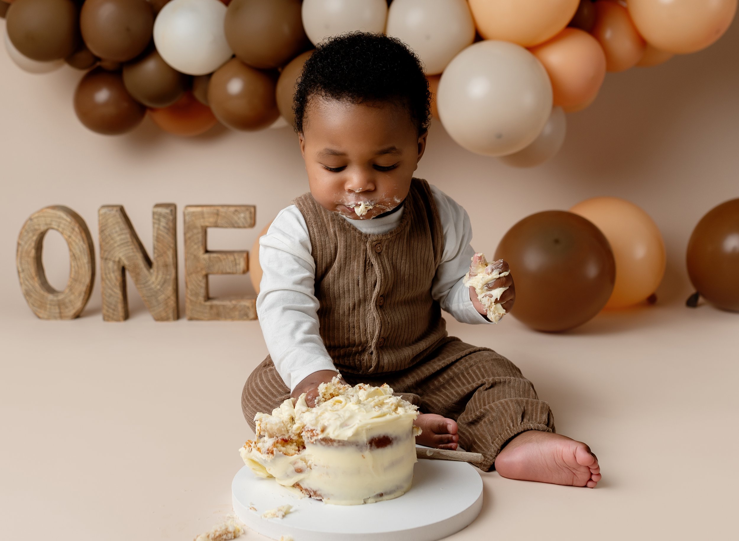 Young child sitting on the floor with a partially eaten birthday cake, surrounded by brown, white, and beige balloons and wooden letters spelling 'ONE' in the background.