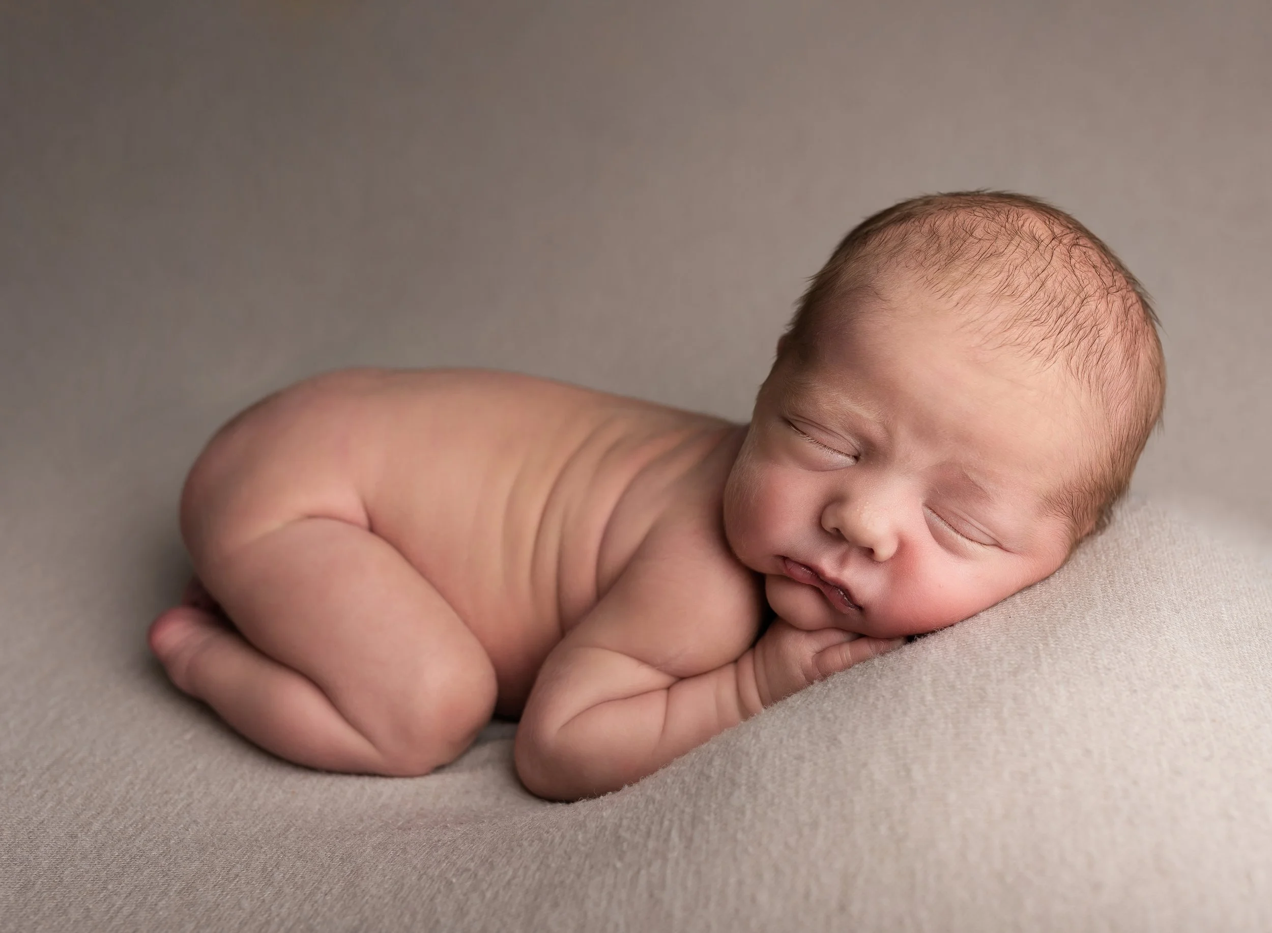 A sleeping newborn baby lying on their side on a soft, neutral-colored surface, with their head resting on their hand and eyes closed.