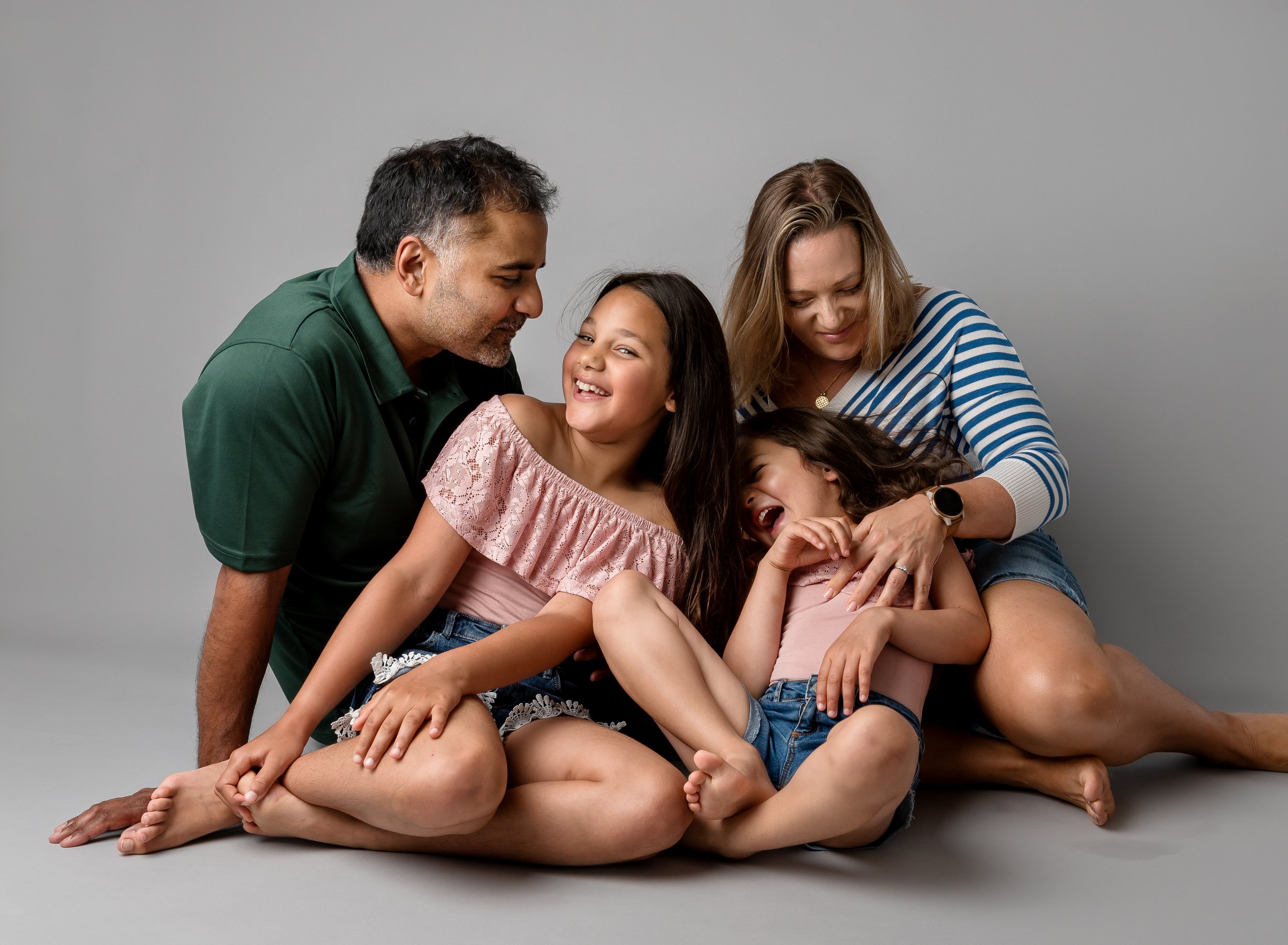 Family of four sitting on the floor, smiling and laughing together against a neutral background.