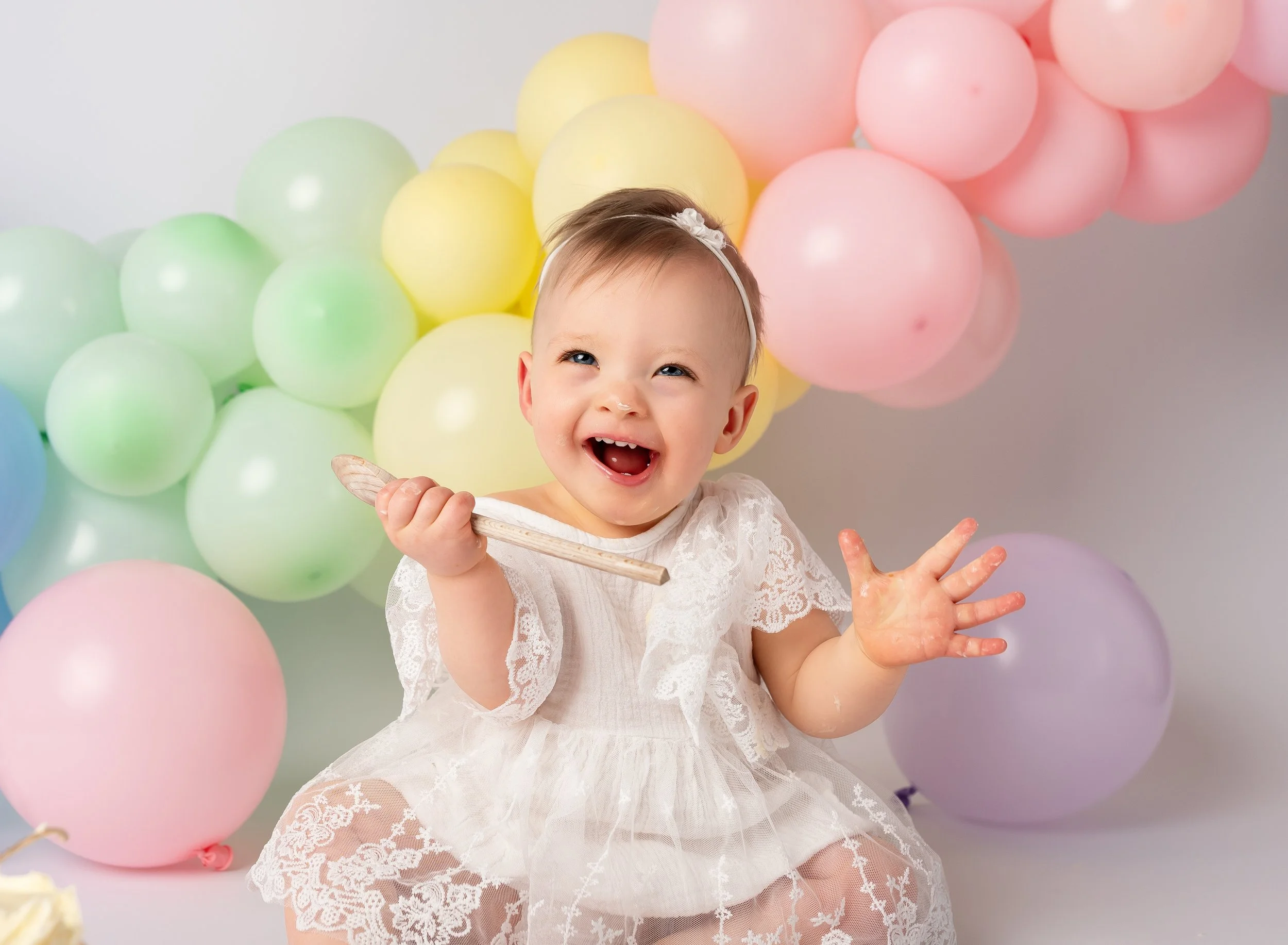 A smiling baby girl in a white lace dress playing with a small wooden toy, surrounded by colorful pastel balloons in yellow, green, pink, purple, and blue.