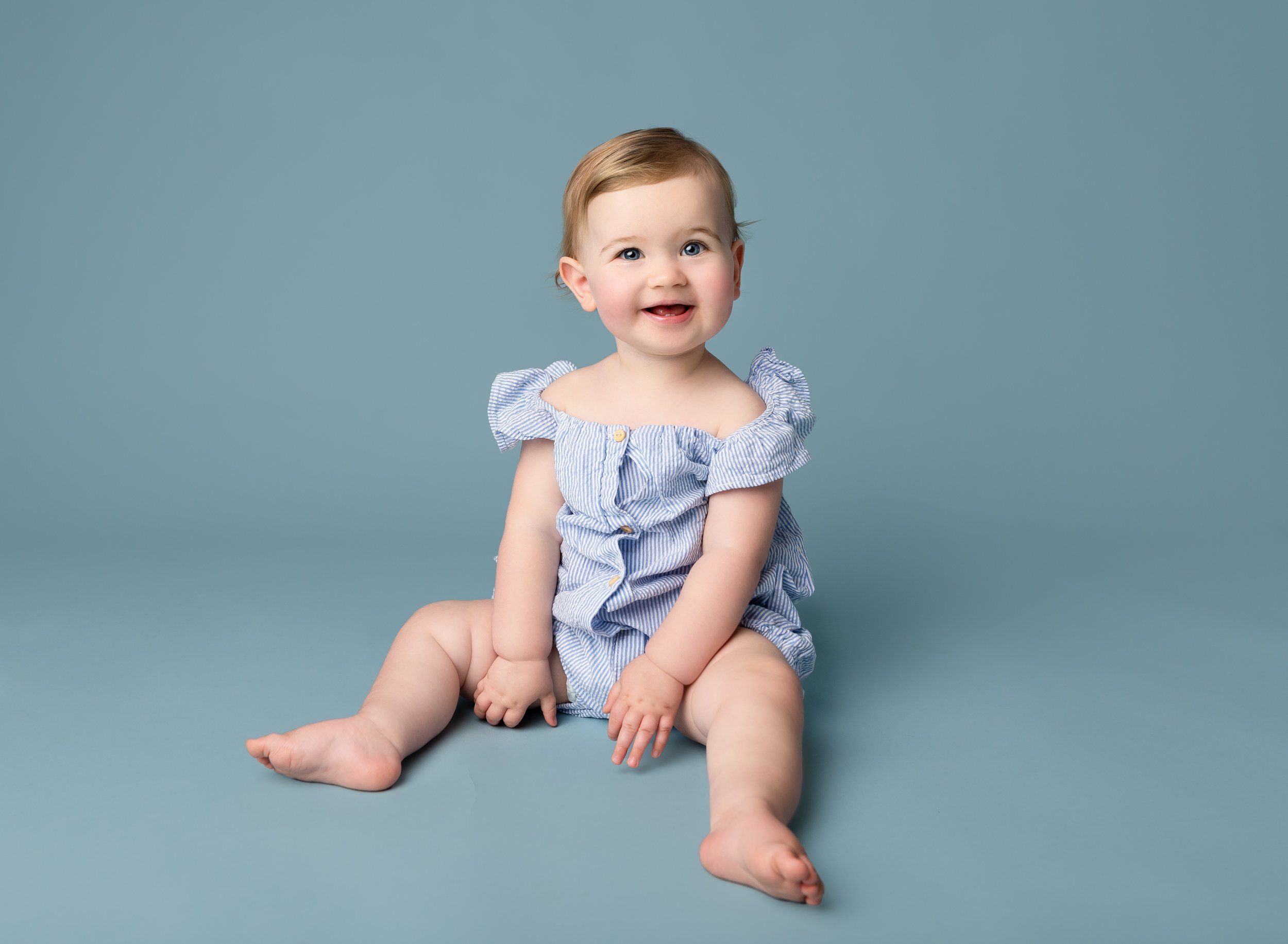 Smiling baby girl with light brown hair and blue eyes sitting on a blue background, wearing a blue and white striped dress with ruffled sleeves.
