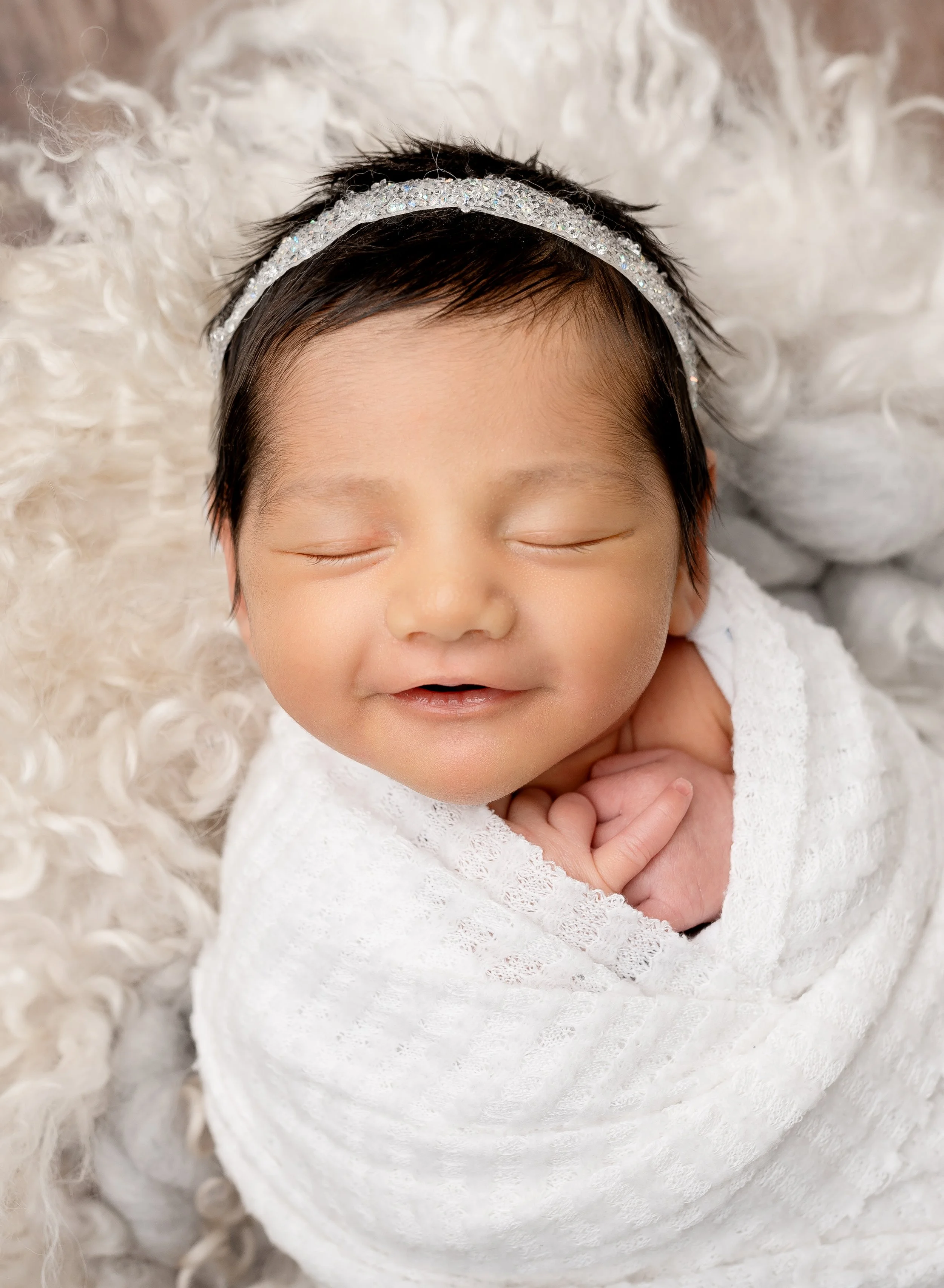 A smiling baby girl with closed eyes, wrapped in a white blanket, wearing a sparkly headband, lying on a fluffy white surface.