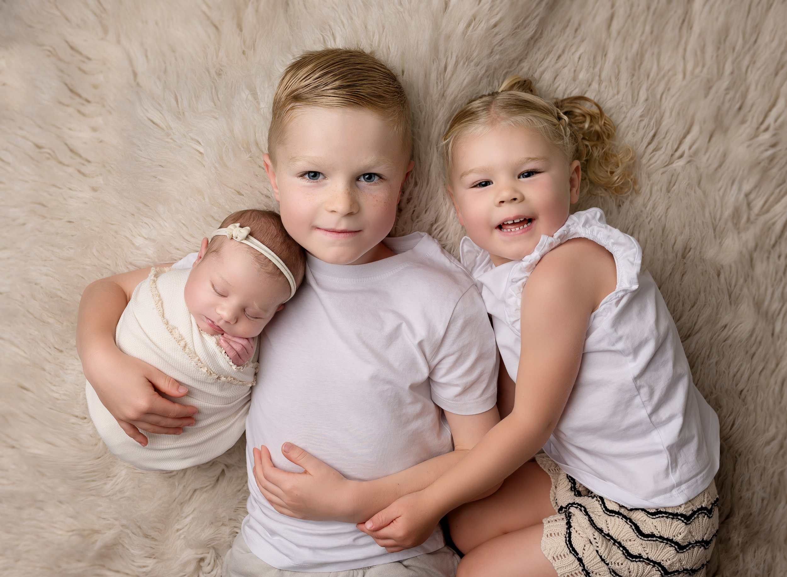 Three children, a girl, a boy, and a baby, lying on a cream-colored furry rug. The baby is sleeping and being held by the boy, who is looking at the camera. The girl is smiling and hugging the boy. The children are wearing white clothing.