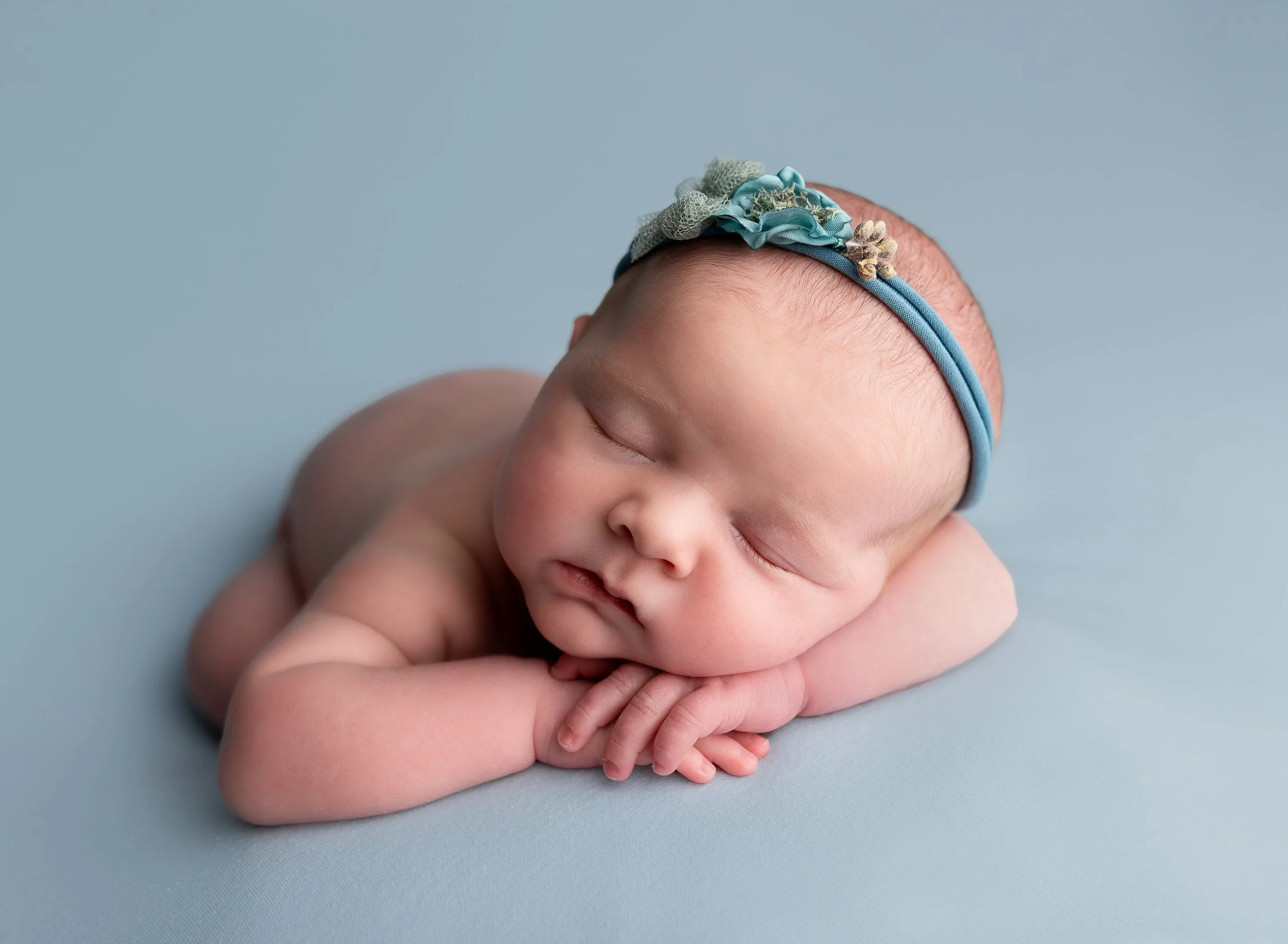Adorable sleeping newborn baby lying on a light blue surface, resting head on crossed arms, wearing a delicate blue headband with fabric flowers.