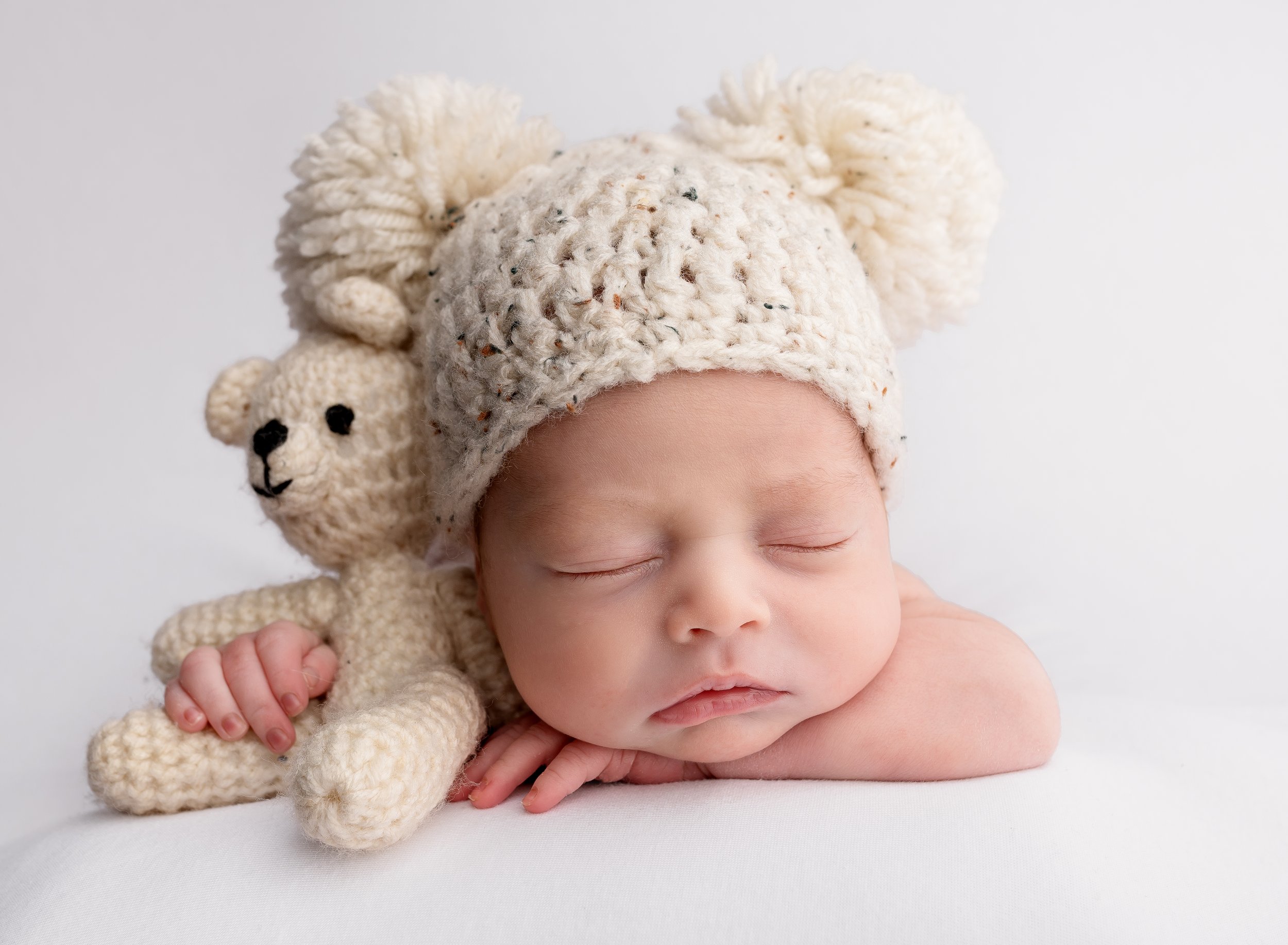 A sleeping baby wearing a beige knitted hat with big pom-poms, resting on a white surface, with a small handmade teddy bear next to its head.