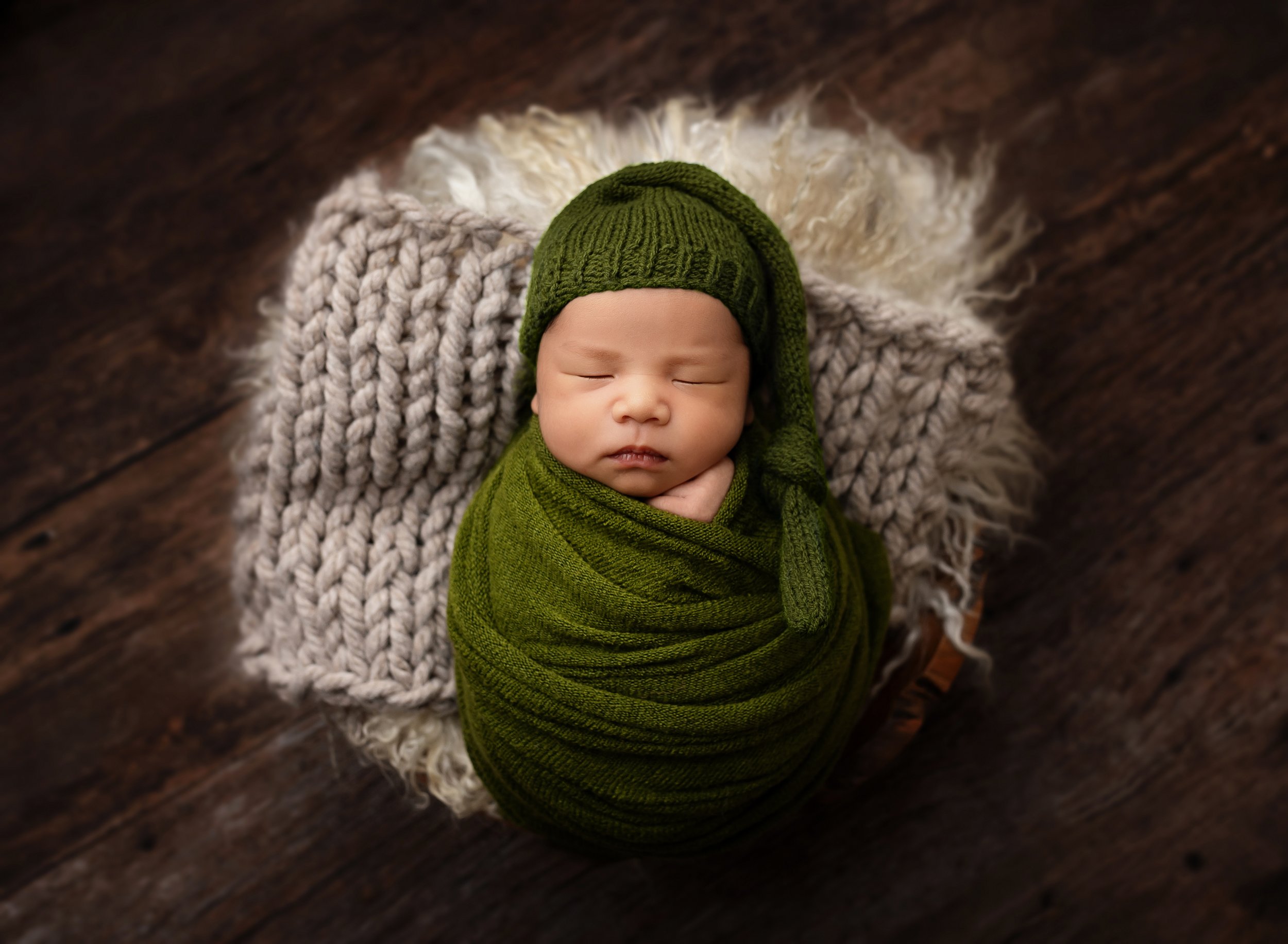 Cute sleeping newborn baby wrapped in a green knitted swaddle and matching green hat, lying on a fluffy cream blanket on a wooden floor.