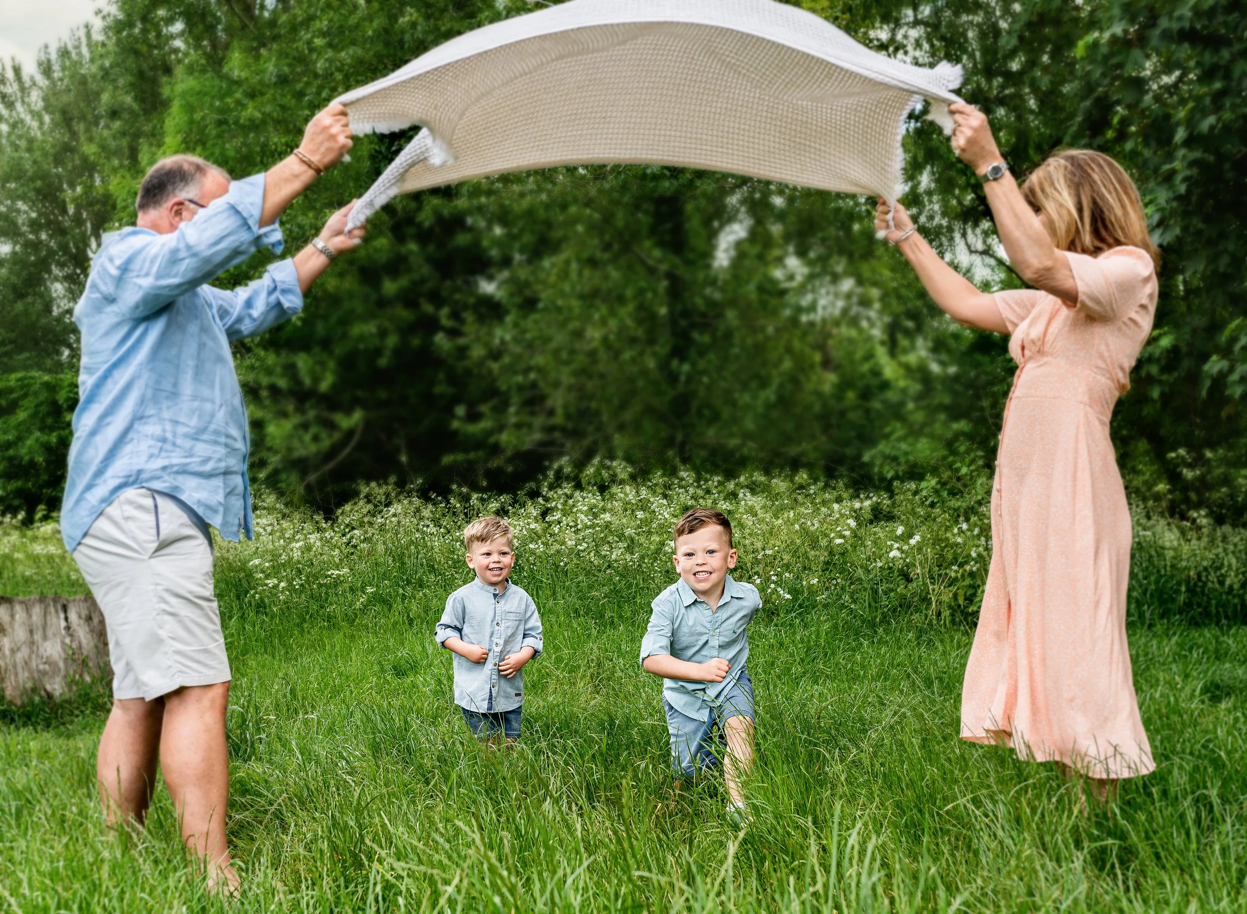 A family with two children playing outside on a grassy field, with grandparents holding a blanket over them as they run underneath, surrounded by green trees.