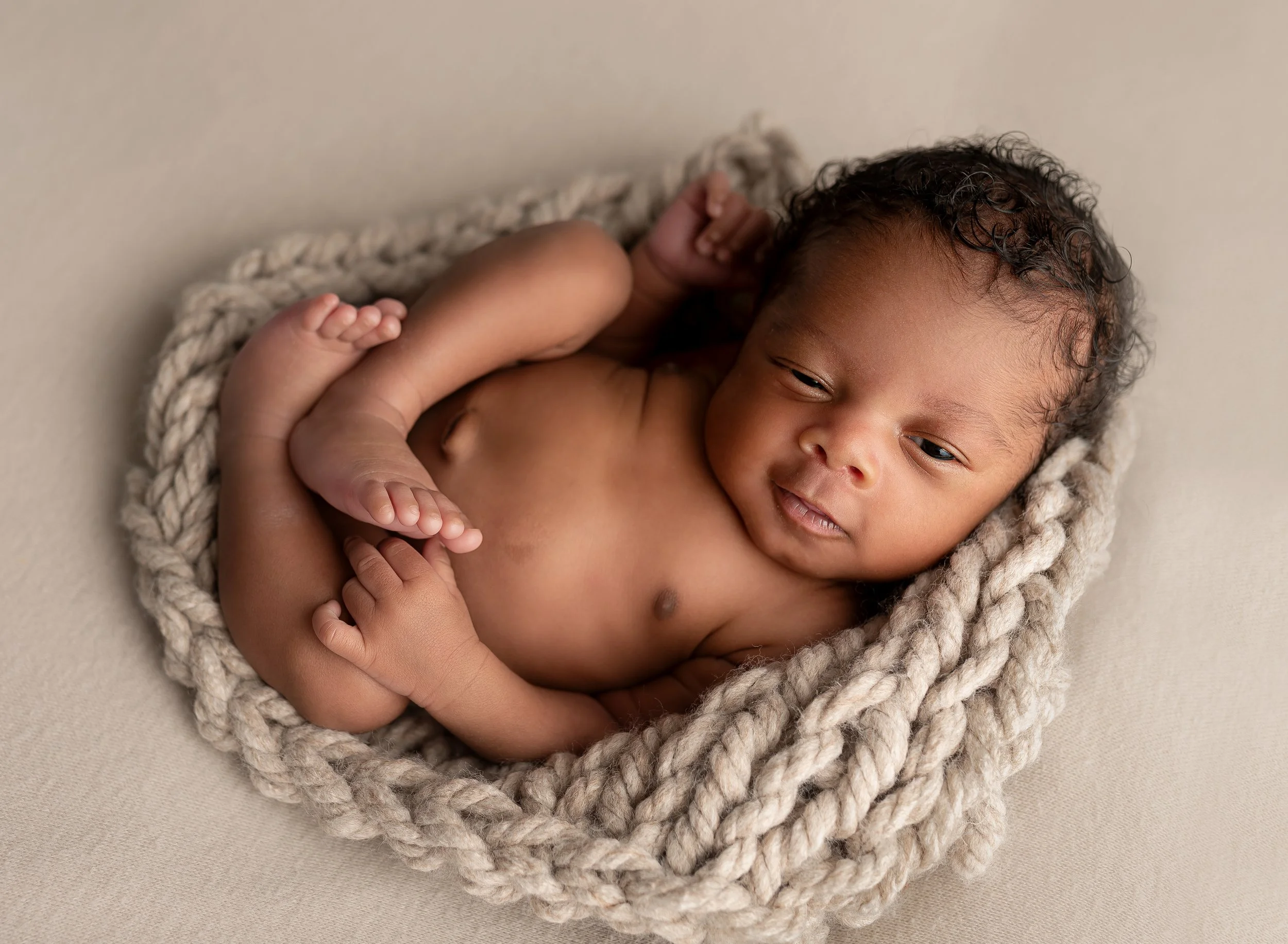 A newborn baby lying on a soft beige surface, nestled inside a chunky knit beige blanket, with a gentle expression and slightly tousled dark hair.