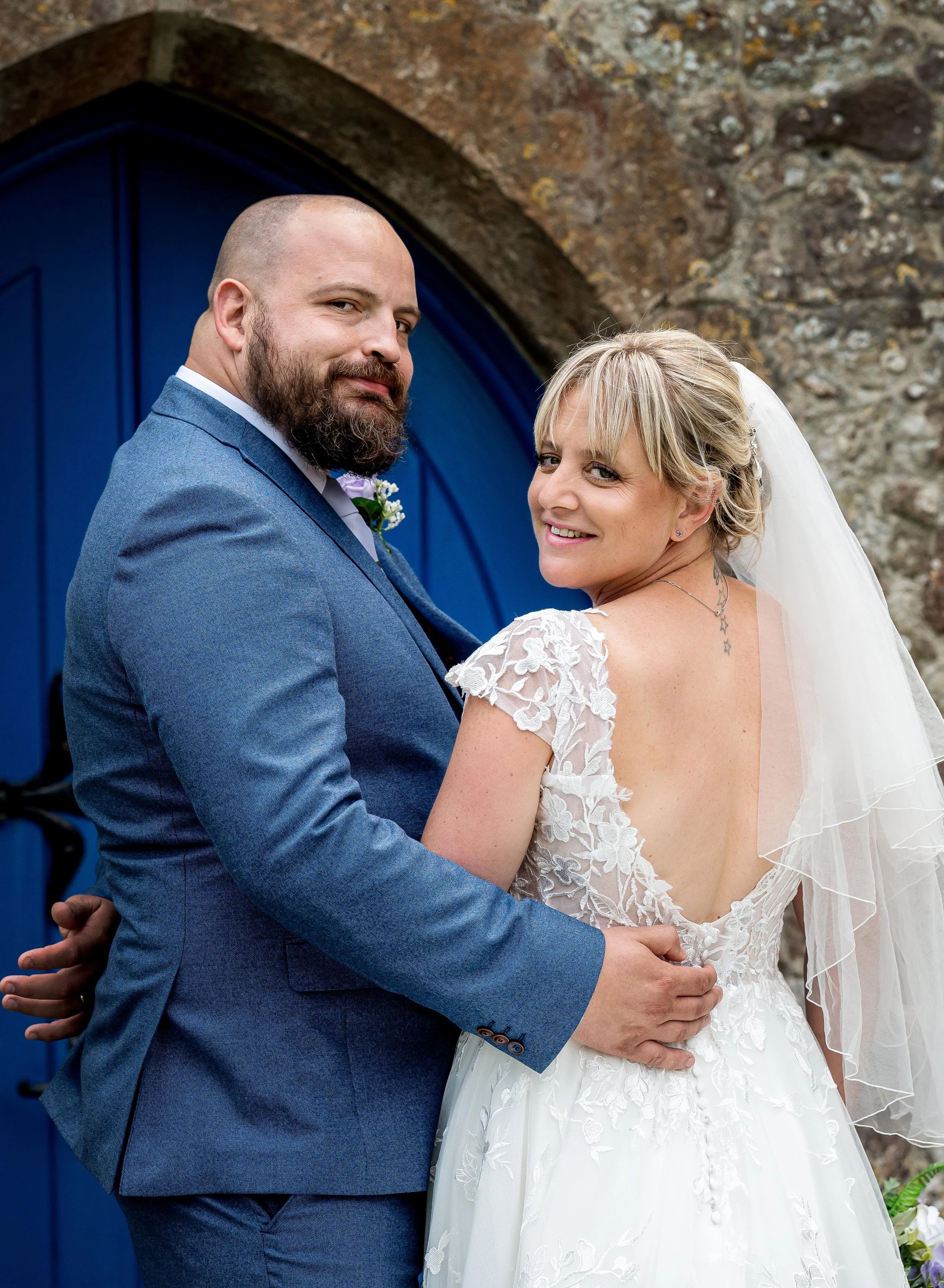 A bride and groom in wedding attire smiling, standing close together outside near a blue door and stone wall.
