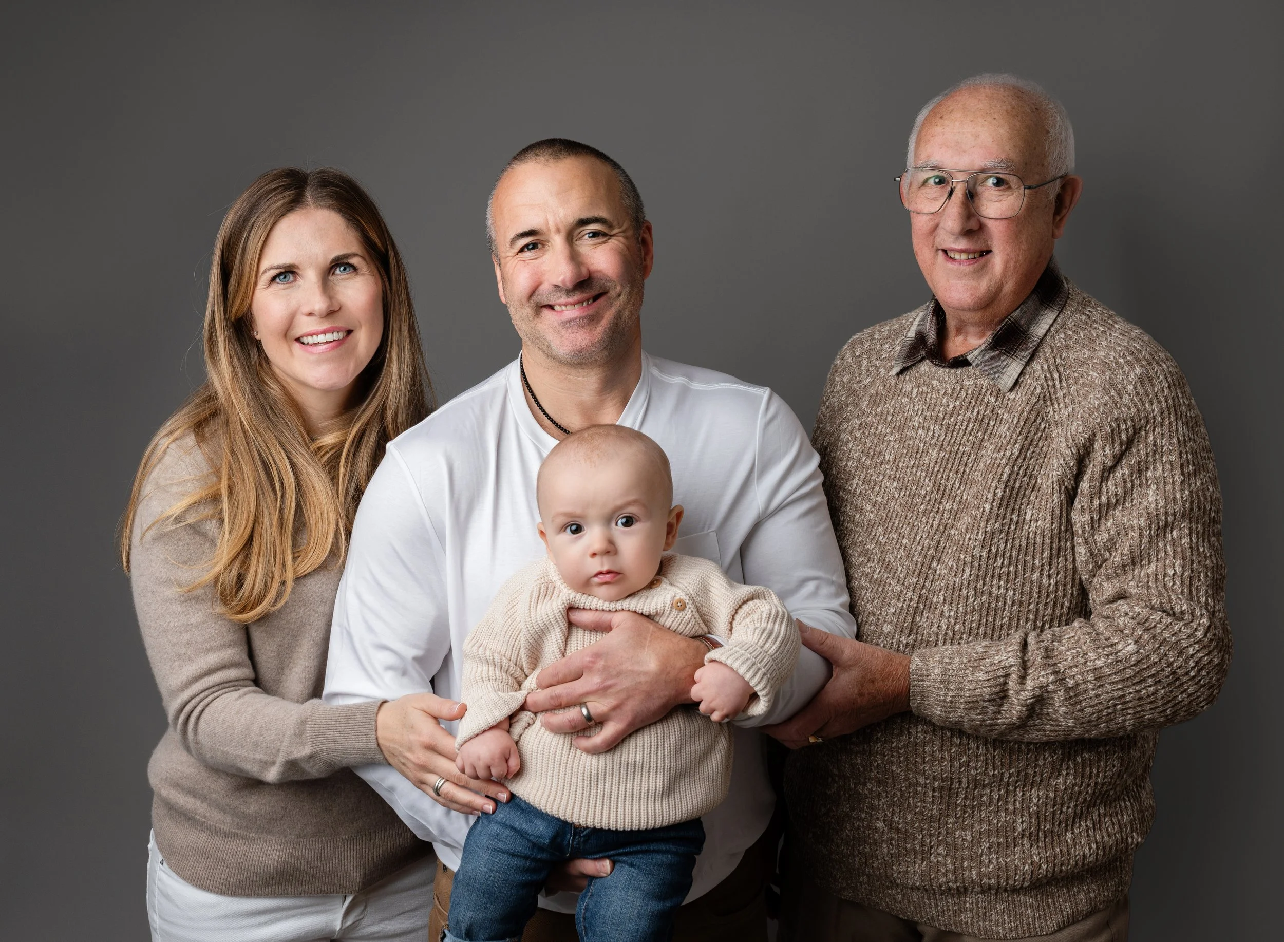 A happy family portrait with a woman, a man, a baby, and an older man, standing together against a gray background.