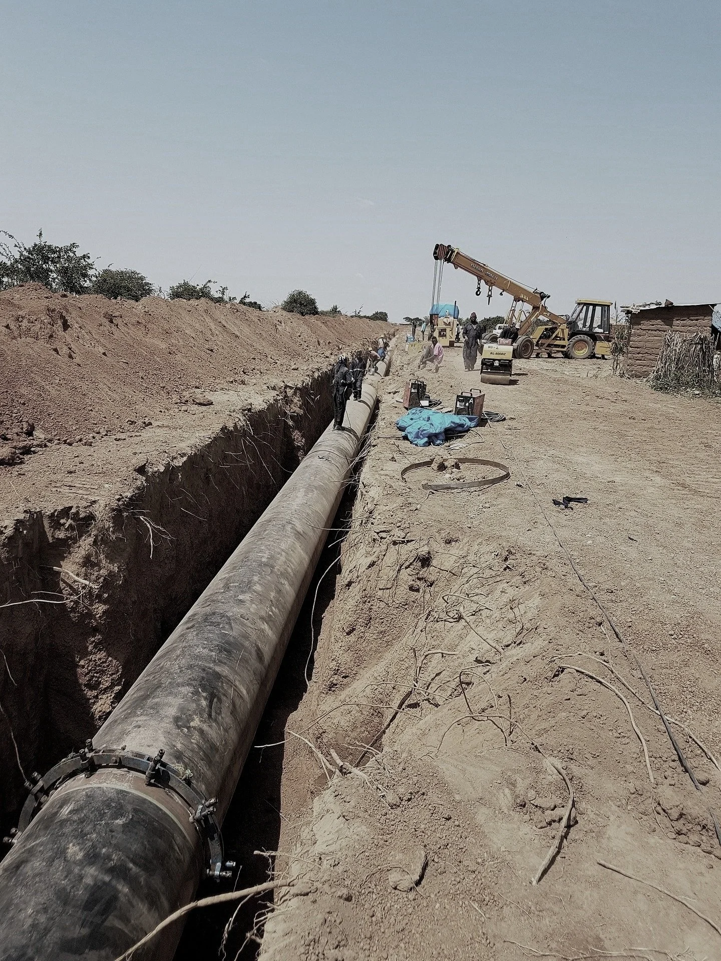 Construction workers installing a large pipeline in a trench in a dry, dusty area with some sparse bushes and a small building in the background.