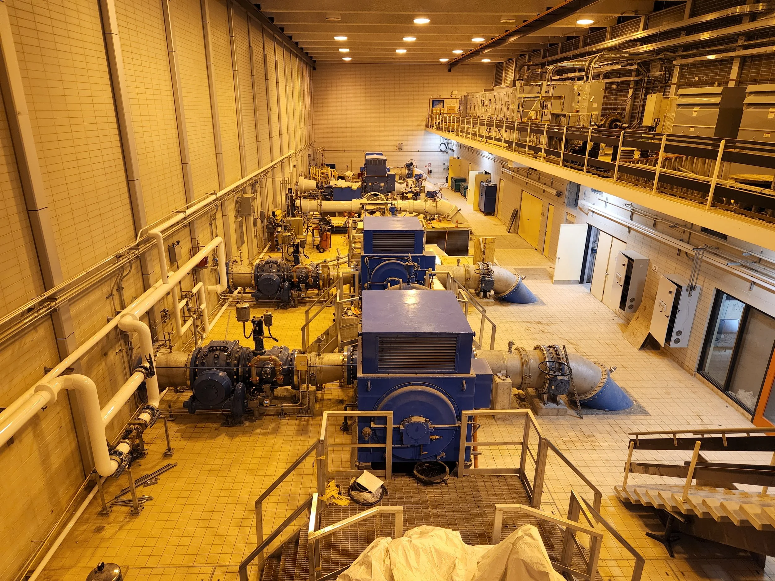 View of an industrial room with large machinery, pipes, and control panels. The room has beige tiled walls and ceiling, with yellow tiled floor, and is well-lit.