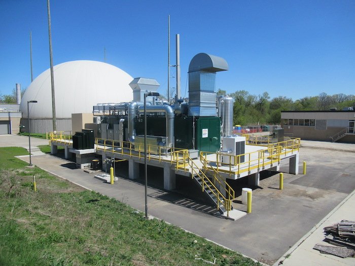 Industrial facility with a green generator or compressor on a raised concrete platform, surrounded by yellow safety railings, pipes, and a large white dome in the background under a clear blue sky.