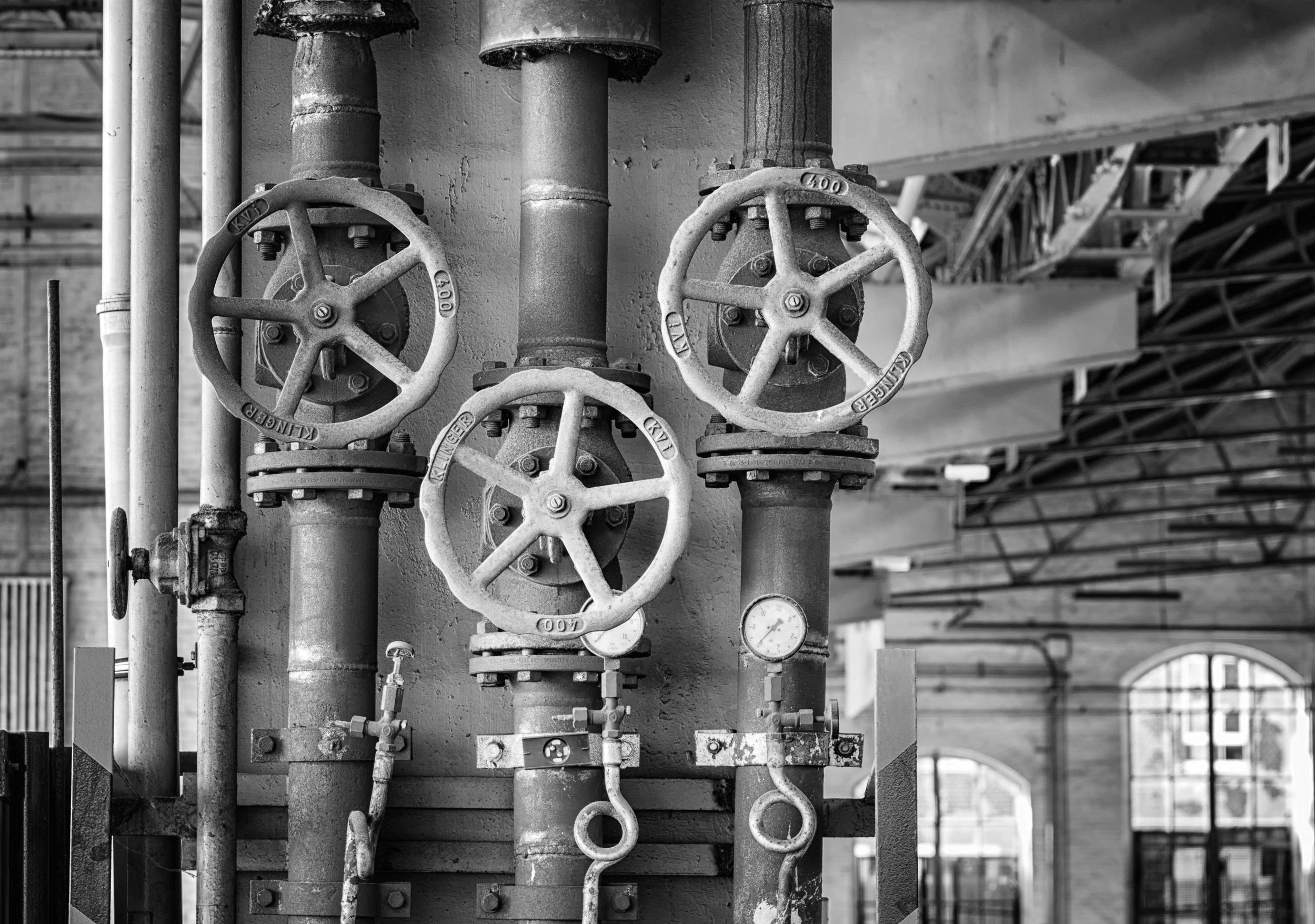 Black and white photo of industrial pipes and valves, with a gauge, in a factory or power plant setting.