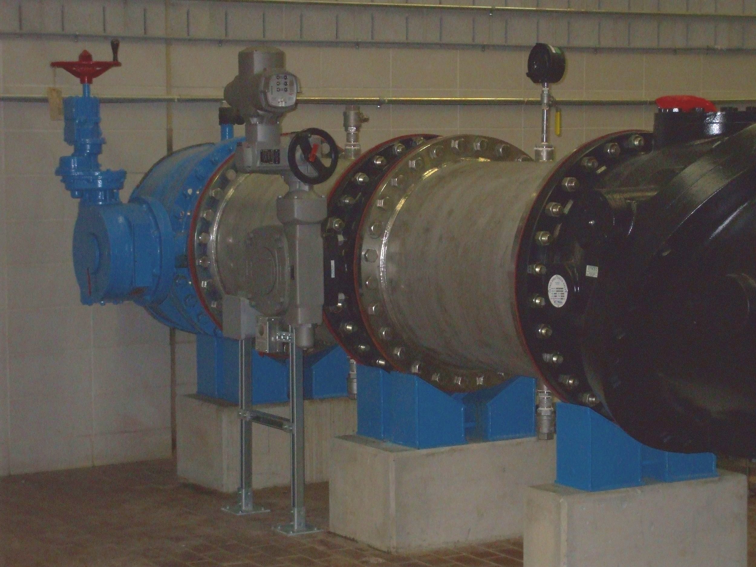 Industrial piping system with large blue valve, pressure gauges, and flanges mounted on a wall with beige tiles.