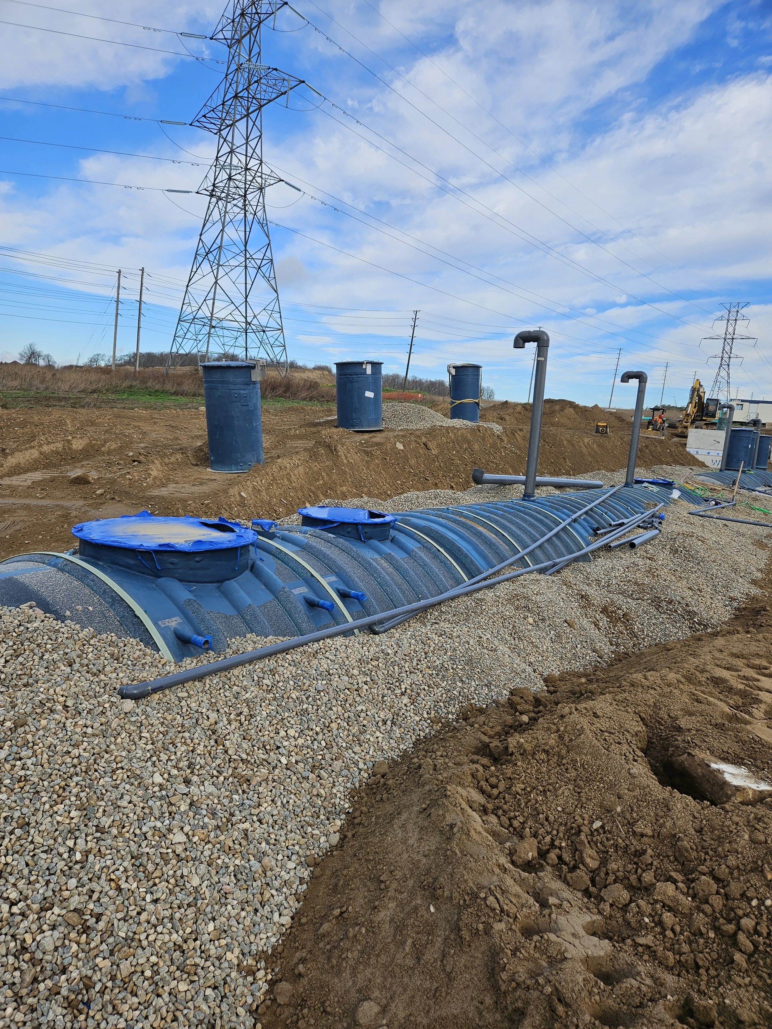 Construction site with large underground pipes, blue barrels, and electrical power lines in the background