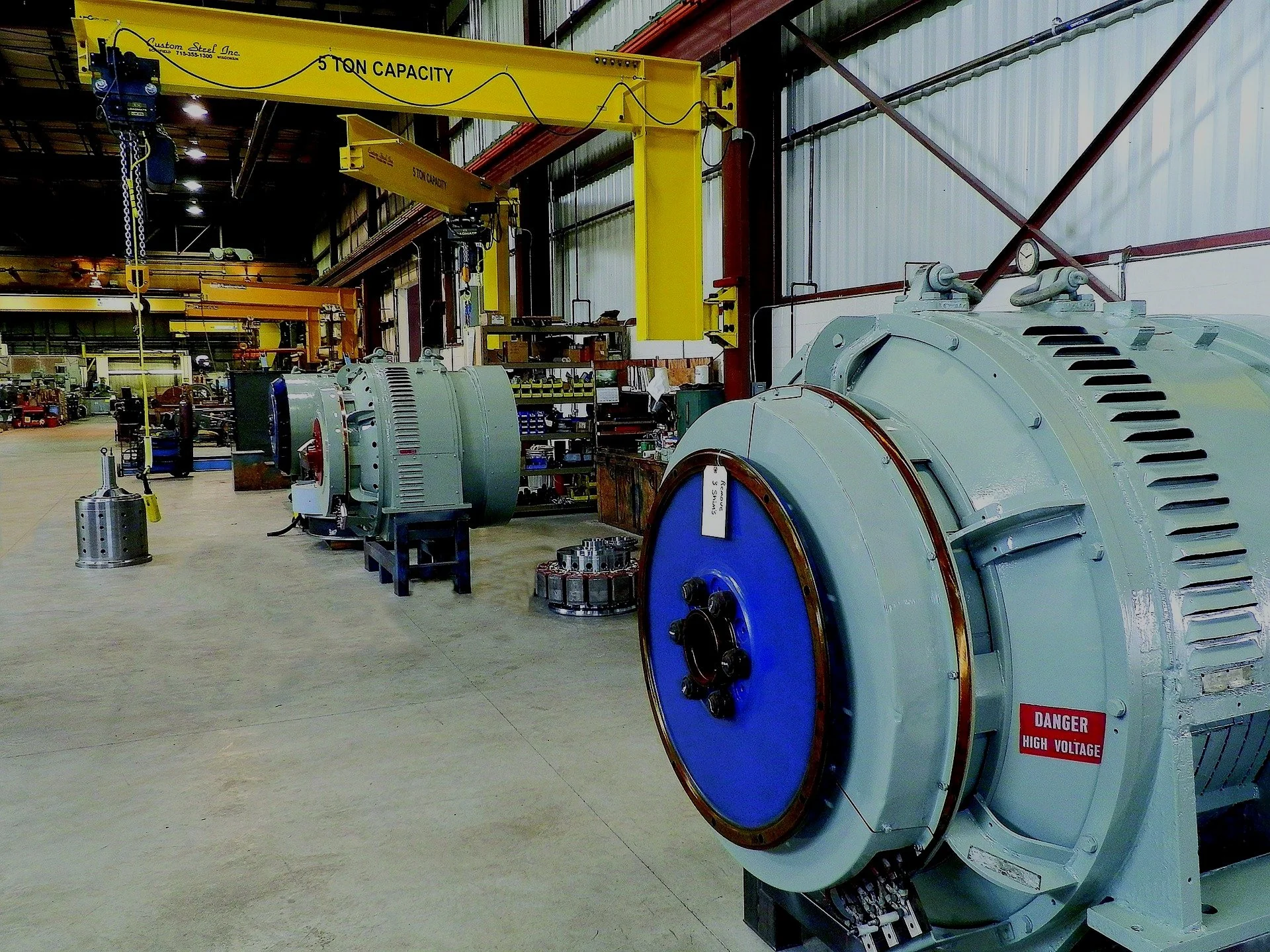 An industrial workshop with large machinery, including a high-voltage generator with a warning label, overhead yellow crane with 5-ton capacity, and various tools and equipment on shelves.