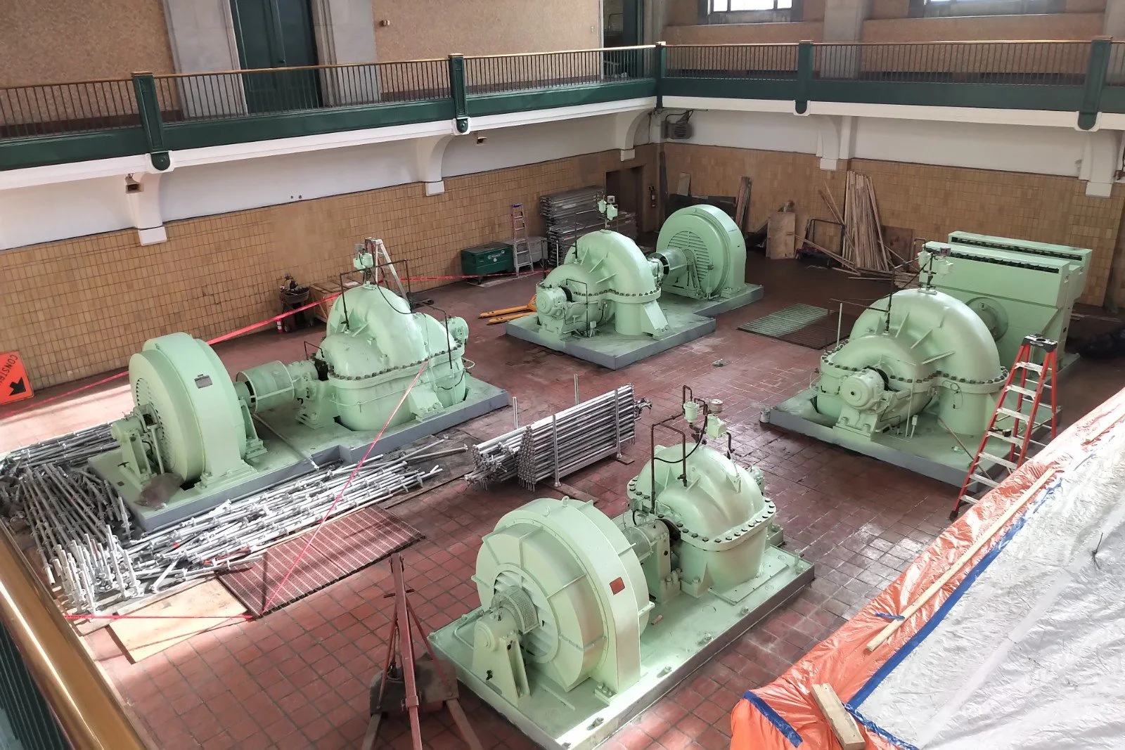 Indoor construction site with six large, light green industrial machines, pipes, and scaffolding on reddish-tiled floor, and a second floor with a railing.