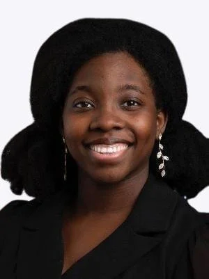 Person smiling, wearing a black outfit and leaf-shaped earrings, with natural hair styled in a large bun on both sides.