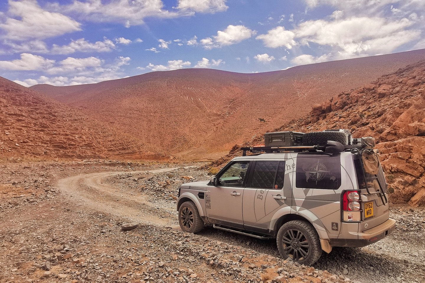 A silver Land Rover off-road vehicle parked on a dirt trail in a rocky, arid mountainous landscape under a partly cloudy sky.