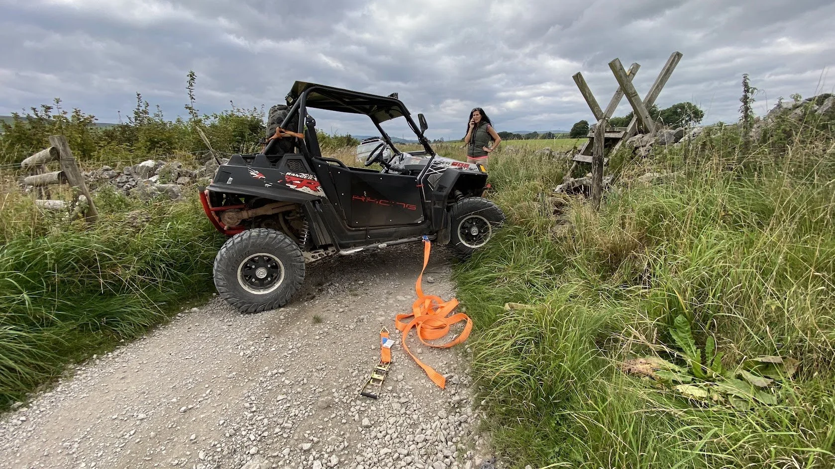 A black off-road utility vehicle is stuck with its front wheels off the ground on a gravel path, surrounded by tall grass and bushes. An orange tow strap is attached to the vehicle, with one end on the ground. A woman stands nearby, looking at the vehicle, with a cloudy sky overhead.