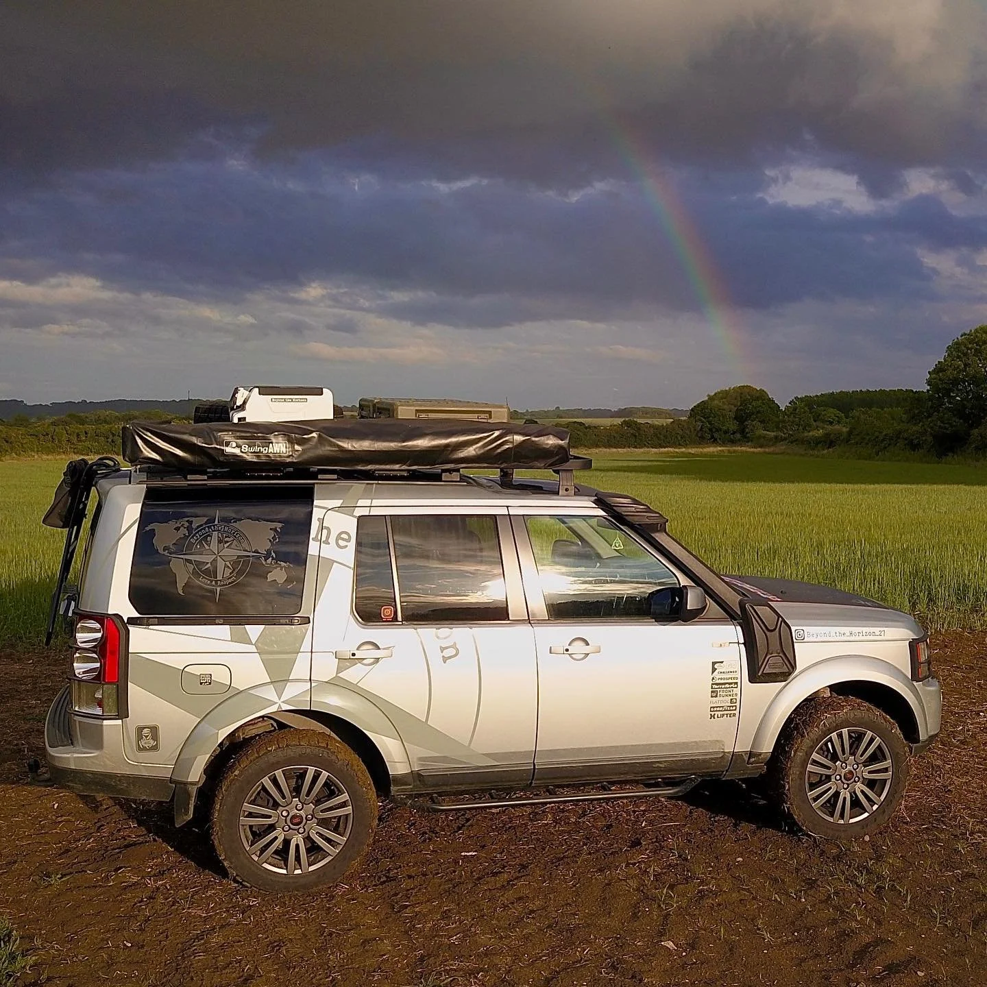 A silver off-road vehicle parked on a dirt field with green grass, with a rainbow in the dark cloudy sky and distant trees and fields in the background.