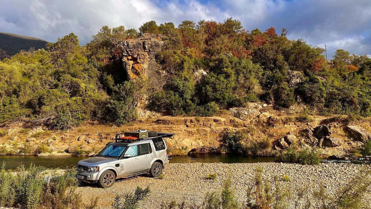 Silver SUV parked on a gravel riverbank with trees, rocks, and a flowing river in the background under a partly cloudy sky.