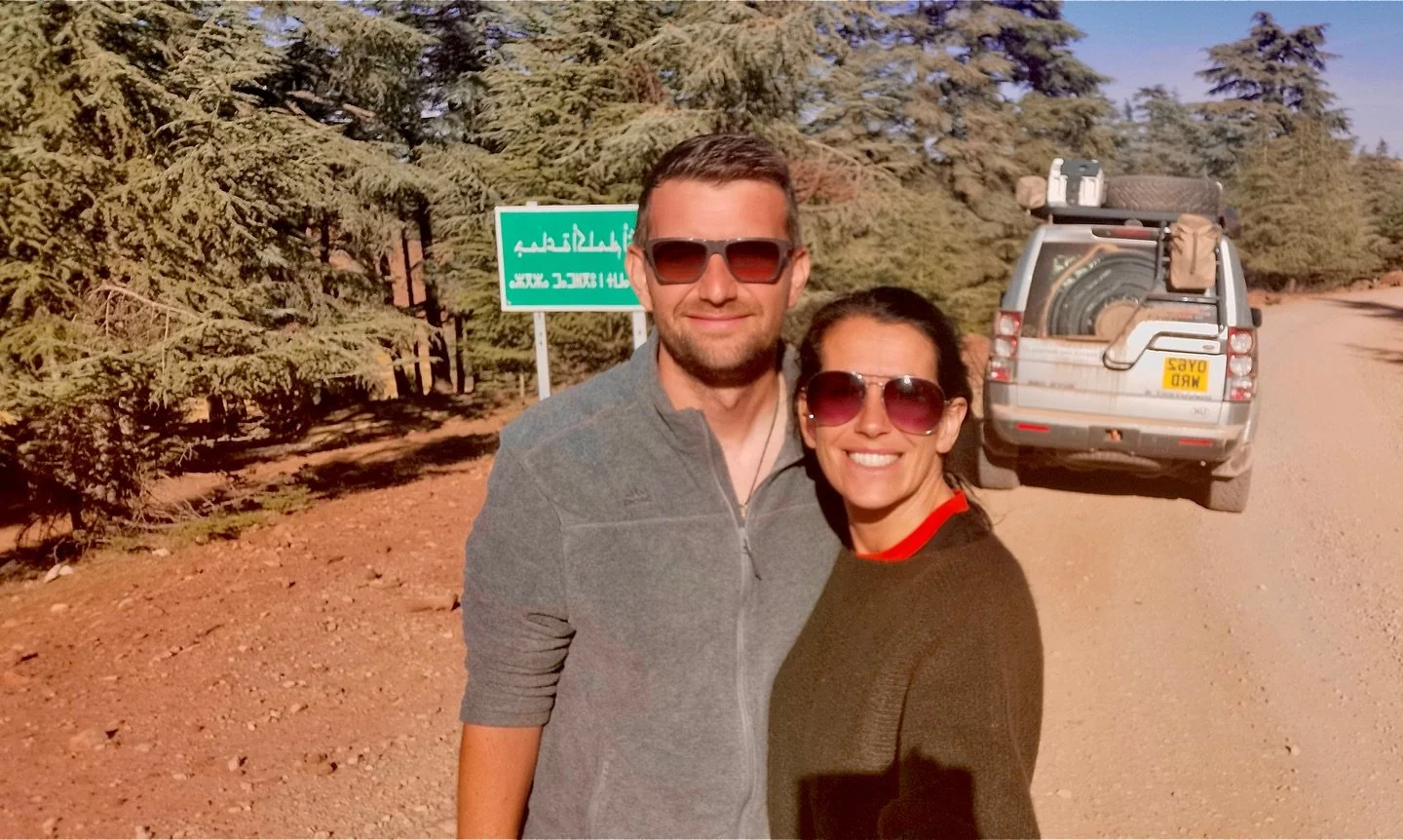 A smiling man and woman wearing sunglasses take a selfie on a dirt road in a desert-like area with trees in the background, and a vehicle loaded with gear behind them.