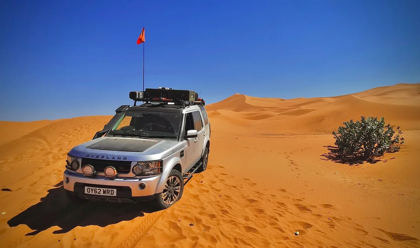 A silver SUV with a flag on top is parked in the sand dunes of a desert under a clear blue sky.