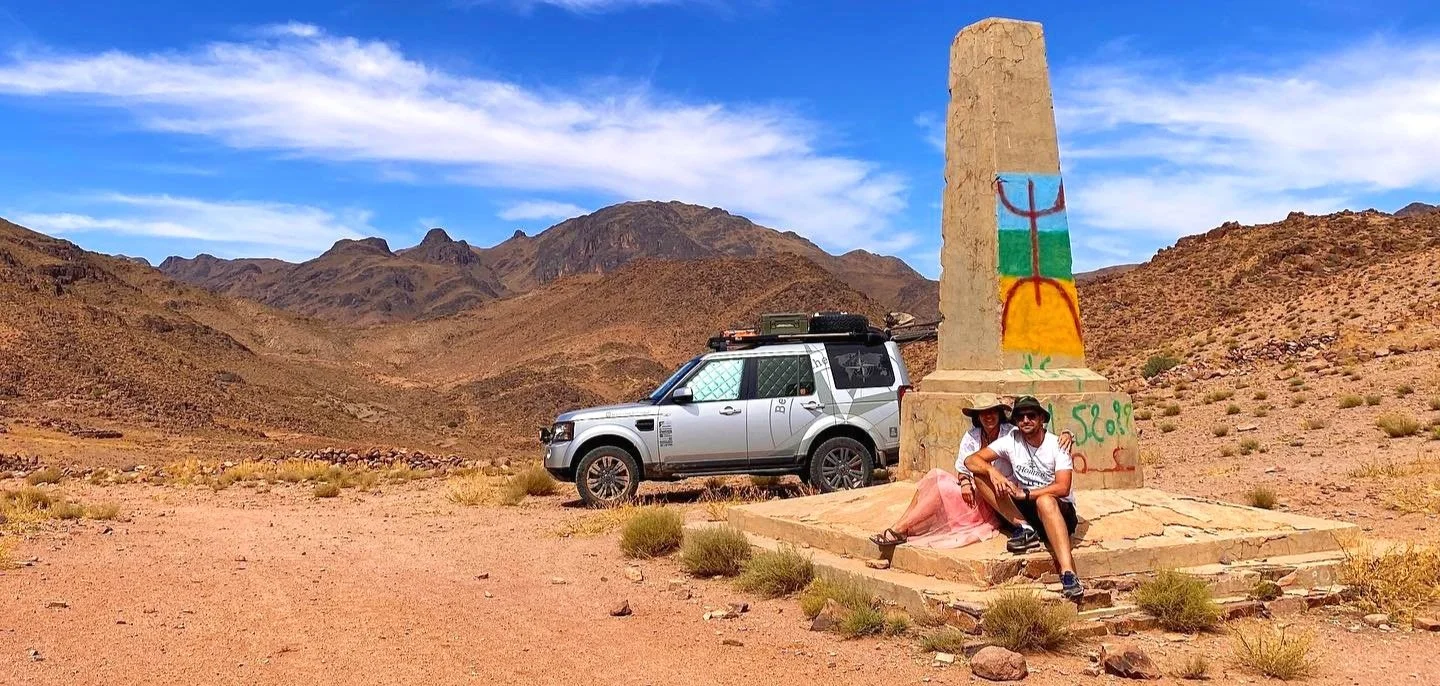 A desert landscape with mountains under a blue sky, featuring a painted obelisk monument, a silver off-road vehicle, and two people sitting at the base of the monument.