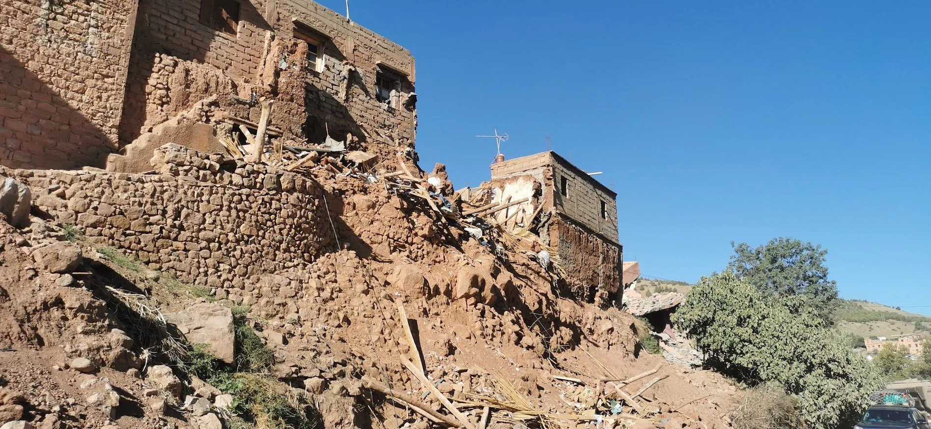 A house on a hillside with significant damage and a landslide, debris scattered on the ground, and a clear blue sky.