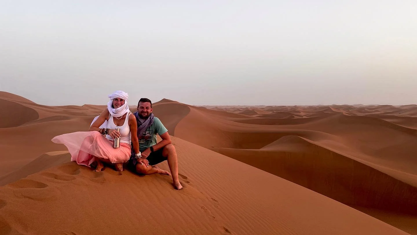Two people sitting on sand dunes in a desert during sunset or sunrise, one woman wearing a white headscarf and long skirt, the other man wearing a scarf and casual clothes.