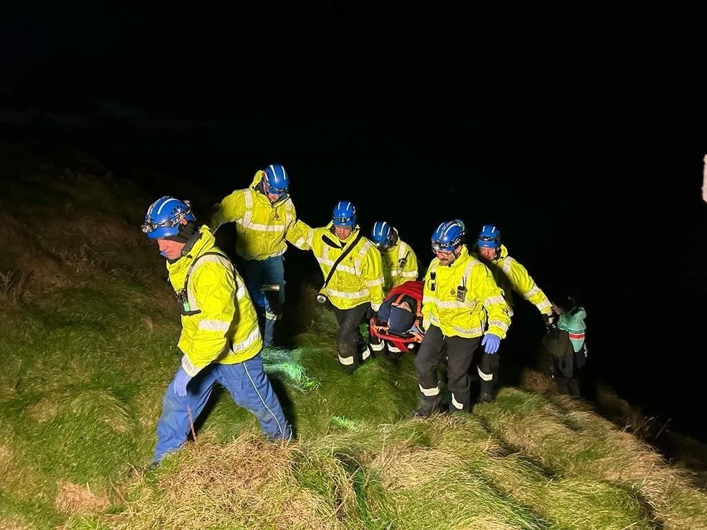 Group of rescue workers in yellow jackets and blue helmets hiking on grassy hillside at night, carrying a stretcher with a person on it.
