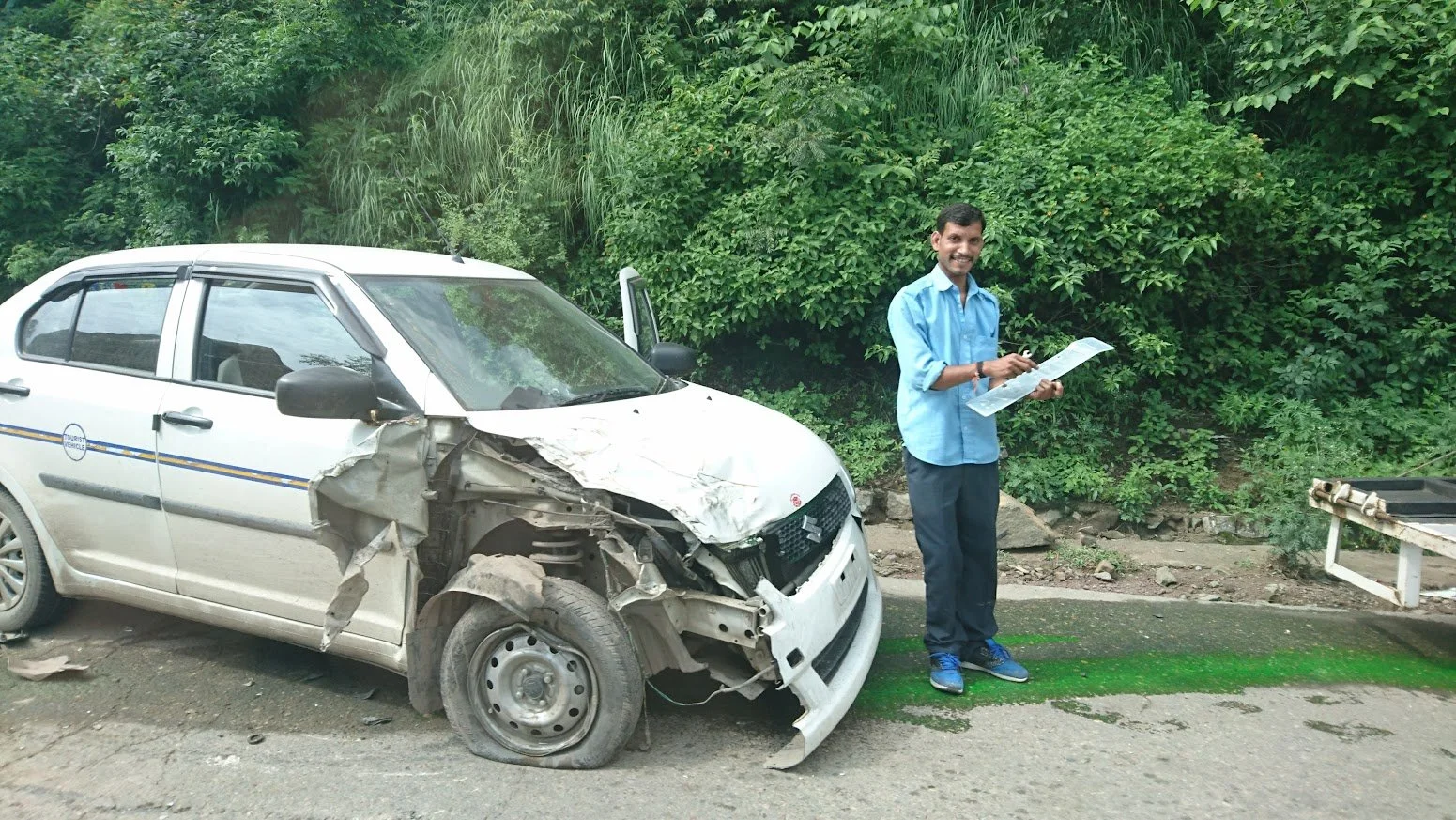 Man in blue shirt standing next to a damaged white car with a crumpled front and missing wheel, on the side of a road surrounded by greenery, holding a paper or document.