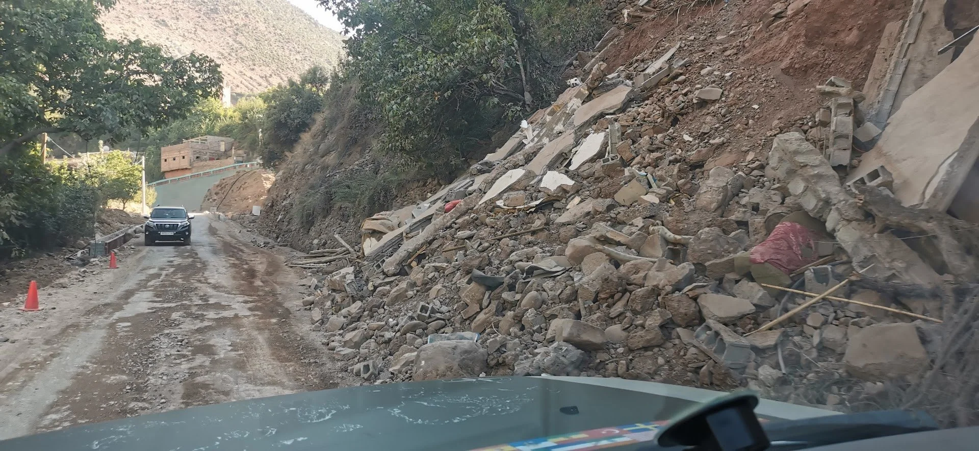 Dirt road with rocks and debris from a landslide, with a black vehicle and orange traffic cones on the road.