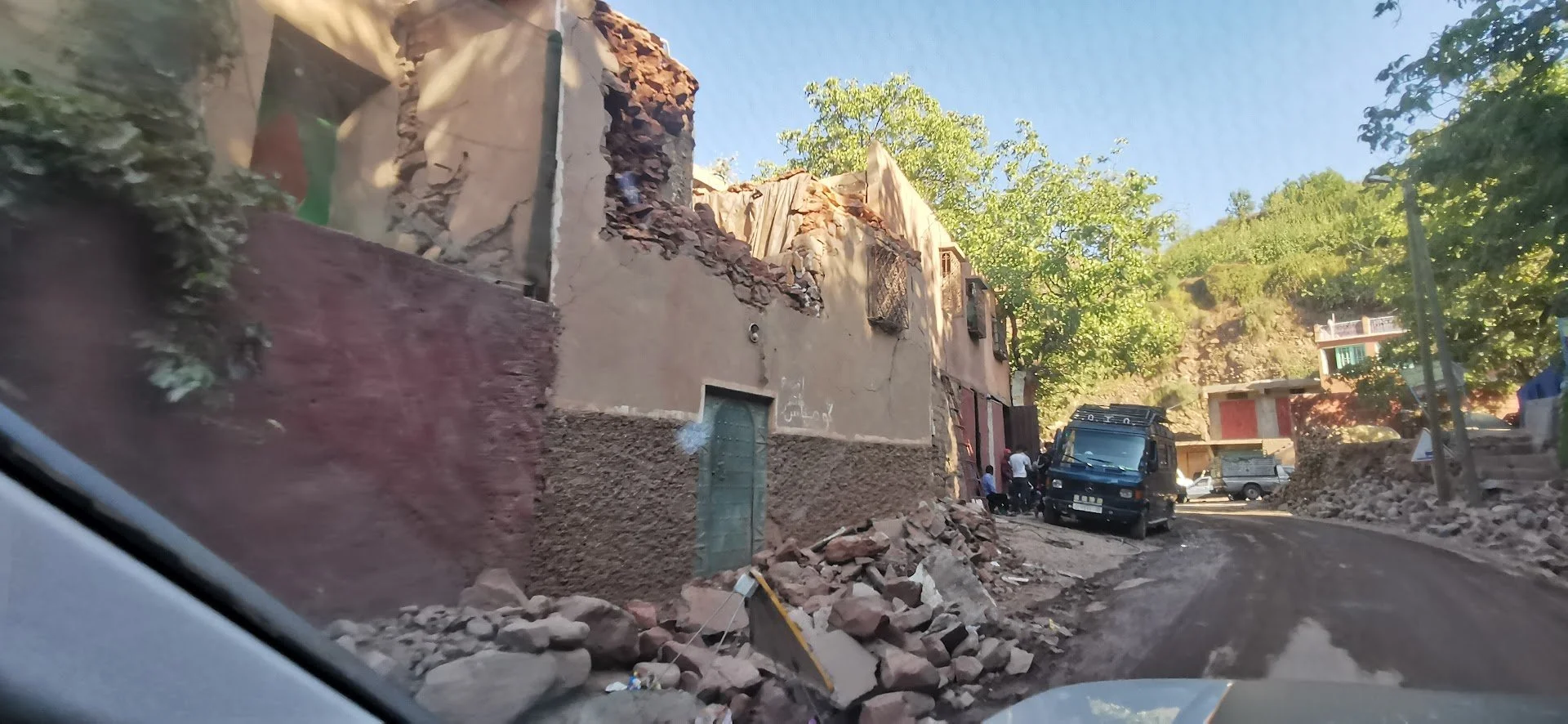 A damaged building with partially collapsed walls and rubble on the ground along a dirt road, with several vehicles and people nearby, in a hilly, wooded area.