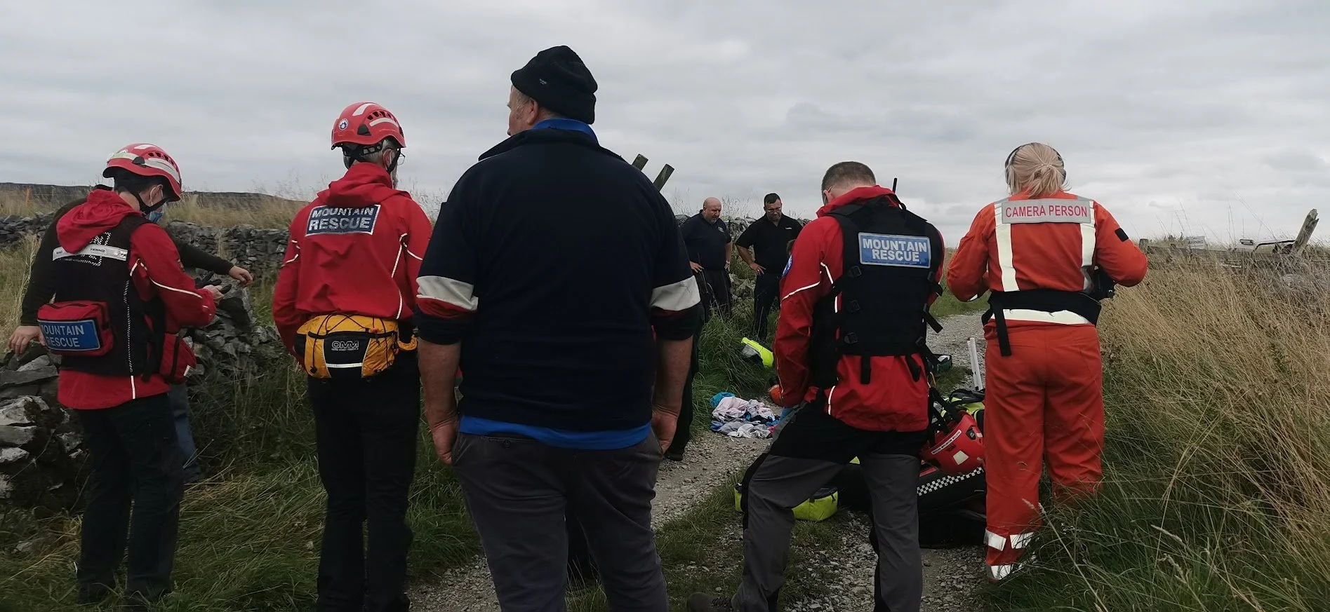 Rescue team members with gear gather outdoors on a cloudy day near a stone wall and grassy terrain.