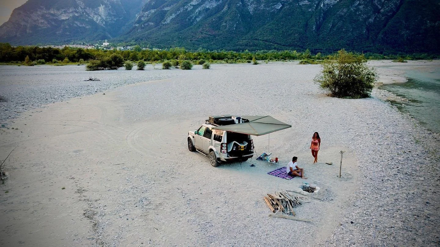 A camping scene on a beach near a river, with a vehicle parked and a canopy setup, two people relaxing and standing nearby, with mountains in the background.