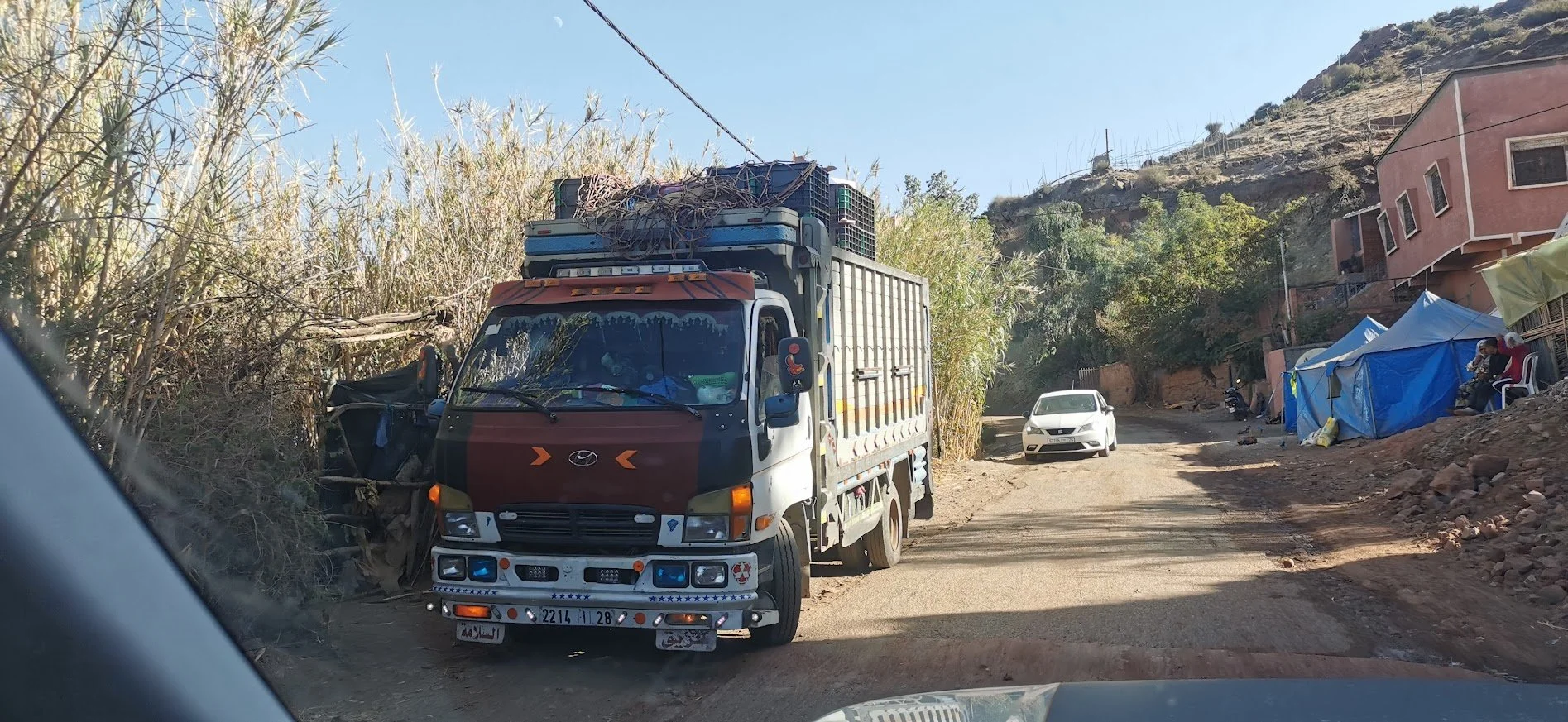 A small truck parked on a dirt road next to makeshift tents and a multi-story building on a hillside.