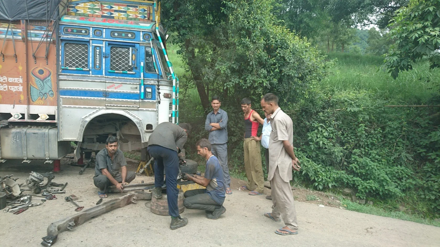 Workers repair a truck on the roadside beside a group of onlookers, with tools and vehicle parts scattered on the ground, and greenery in the background.